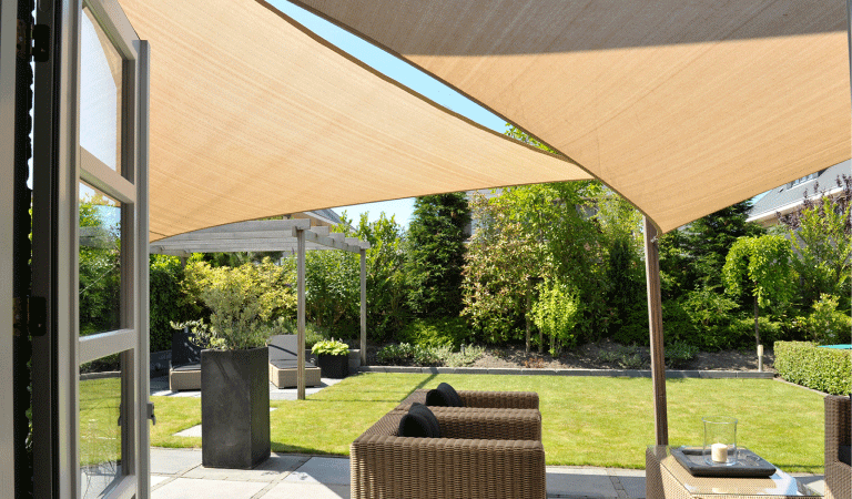 Zicht op tuin vanuit openslaande deur met op de voorgrond een zithoek, overdekt met schaduwdoeken. Op de achtergrond een gazon en pergola.