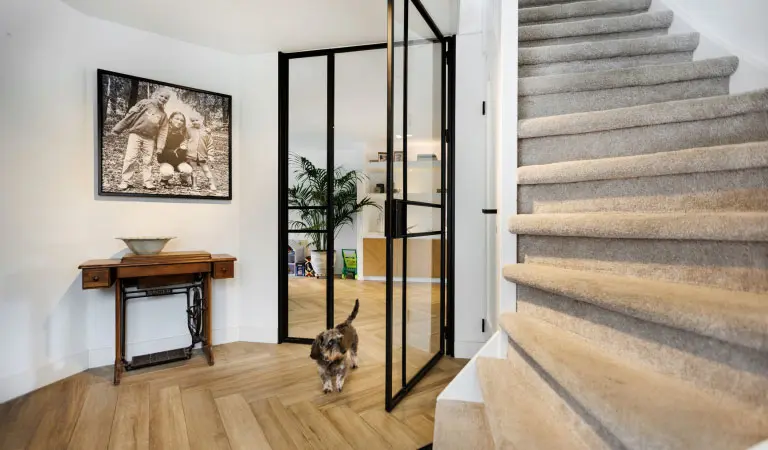 Ruime entree met visgraatparketvloer, taatsdeur en gedraaide trap met beige tapijt. In de hal staat een kat.