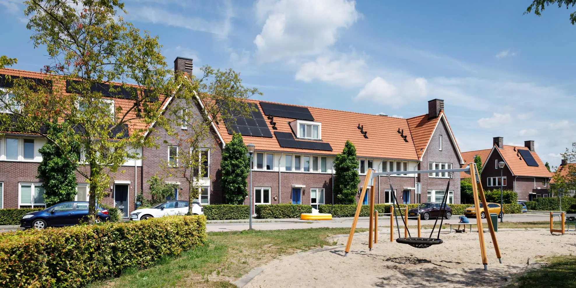 Rijtjeshuizen met op de voorgrond een kinderspeelplaats met schommel en bosjes.