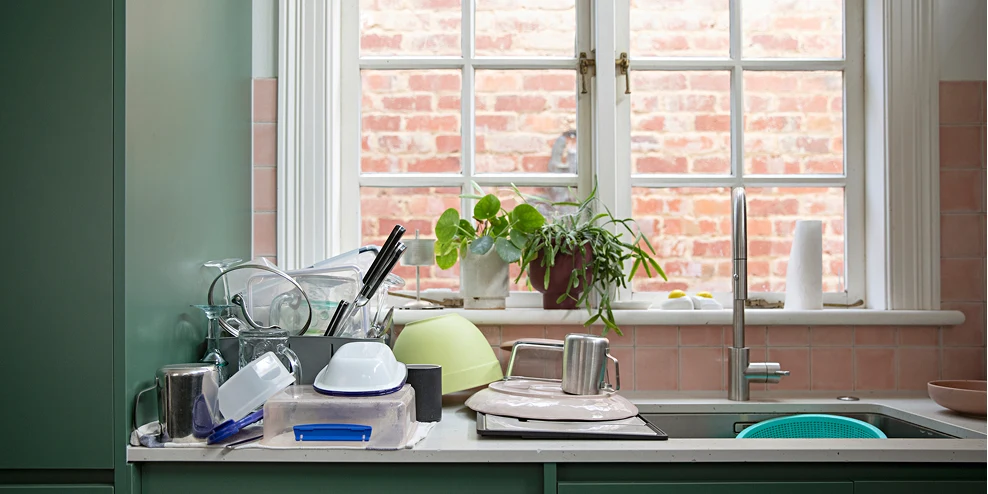 Rommelig aanrechtblad vol afwas en vuile vaat voor keukenraam met uitzicht op muur.