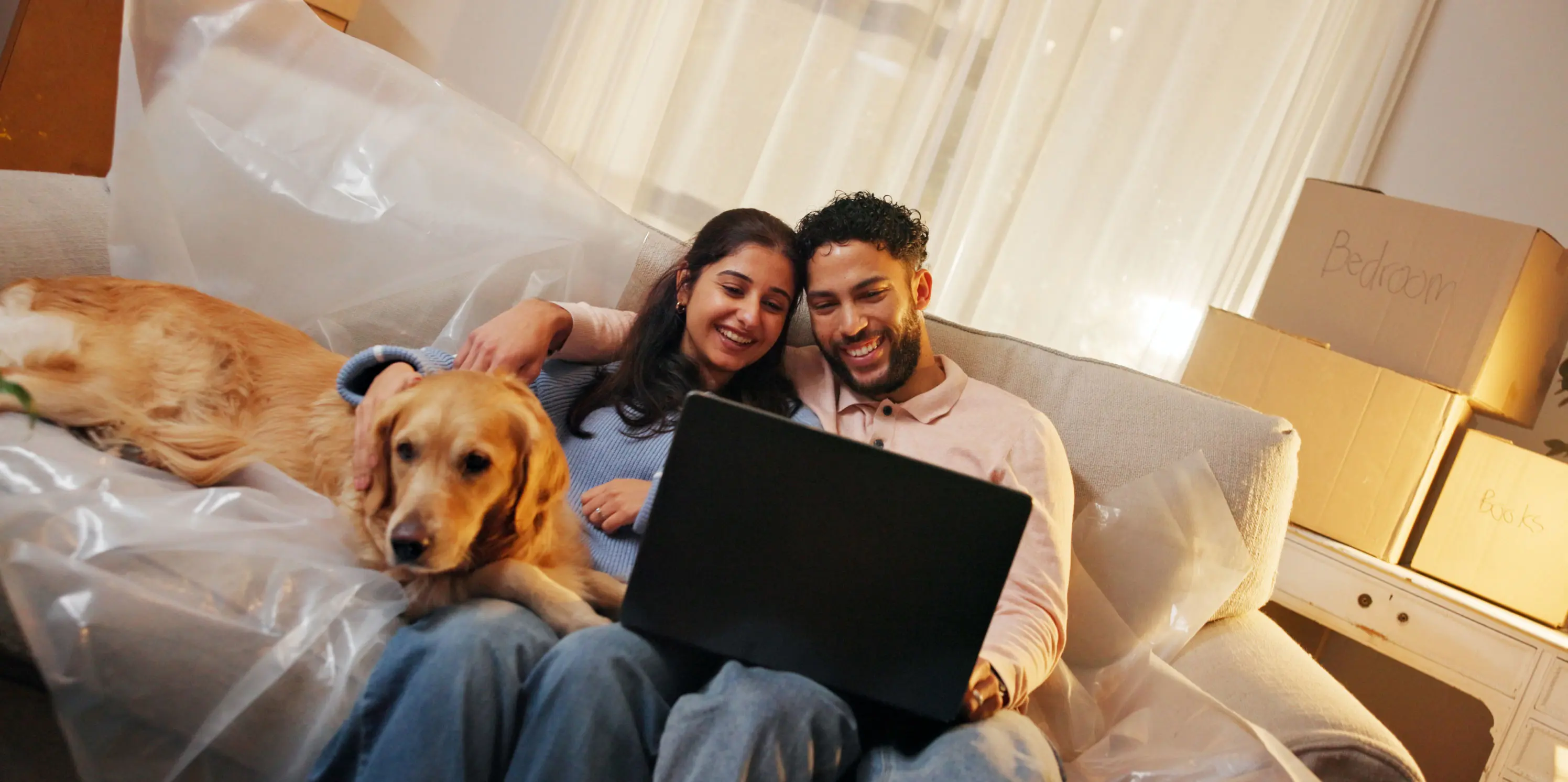Stel zit met laptop en schoot en hond naast zich op een bank die nog half in plastic is verpakt, met op de achtergrond verhuisdozen. 