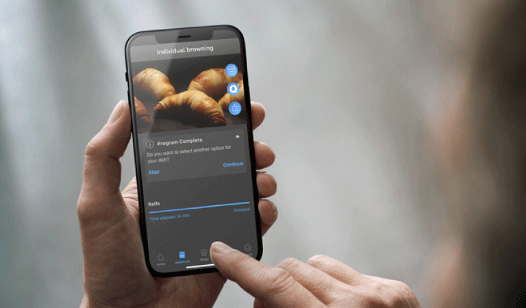 Hand houdt smartphone vast waarop een ovenprogramma gekozen kan worden voor de juiste bereiding van croissants. 