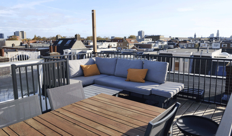 Blik op het dakterras van het huis, met daarop een blauwe loungebank en een houten eettafel. 