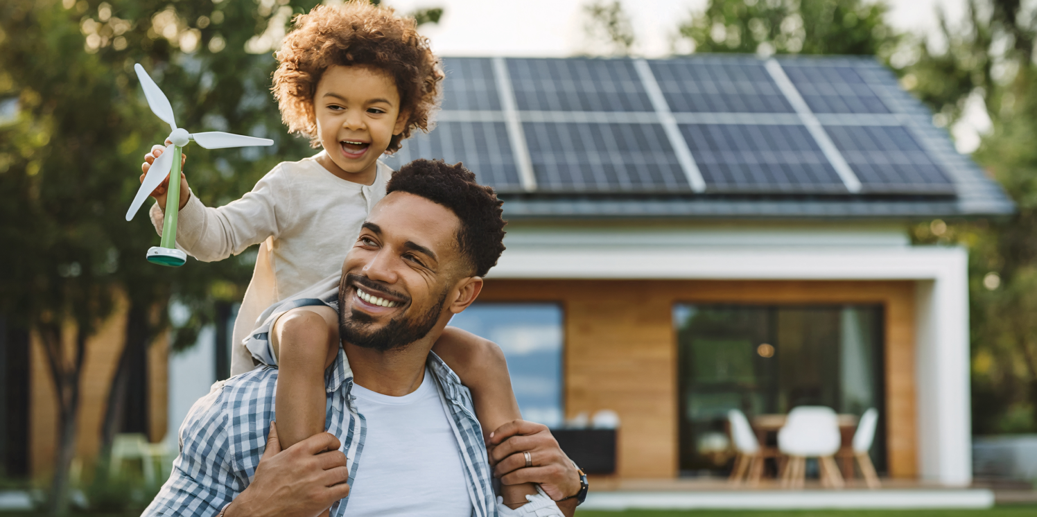 Vader met dochtertje op zijn schouders voor een huis met zonnepanelen.