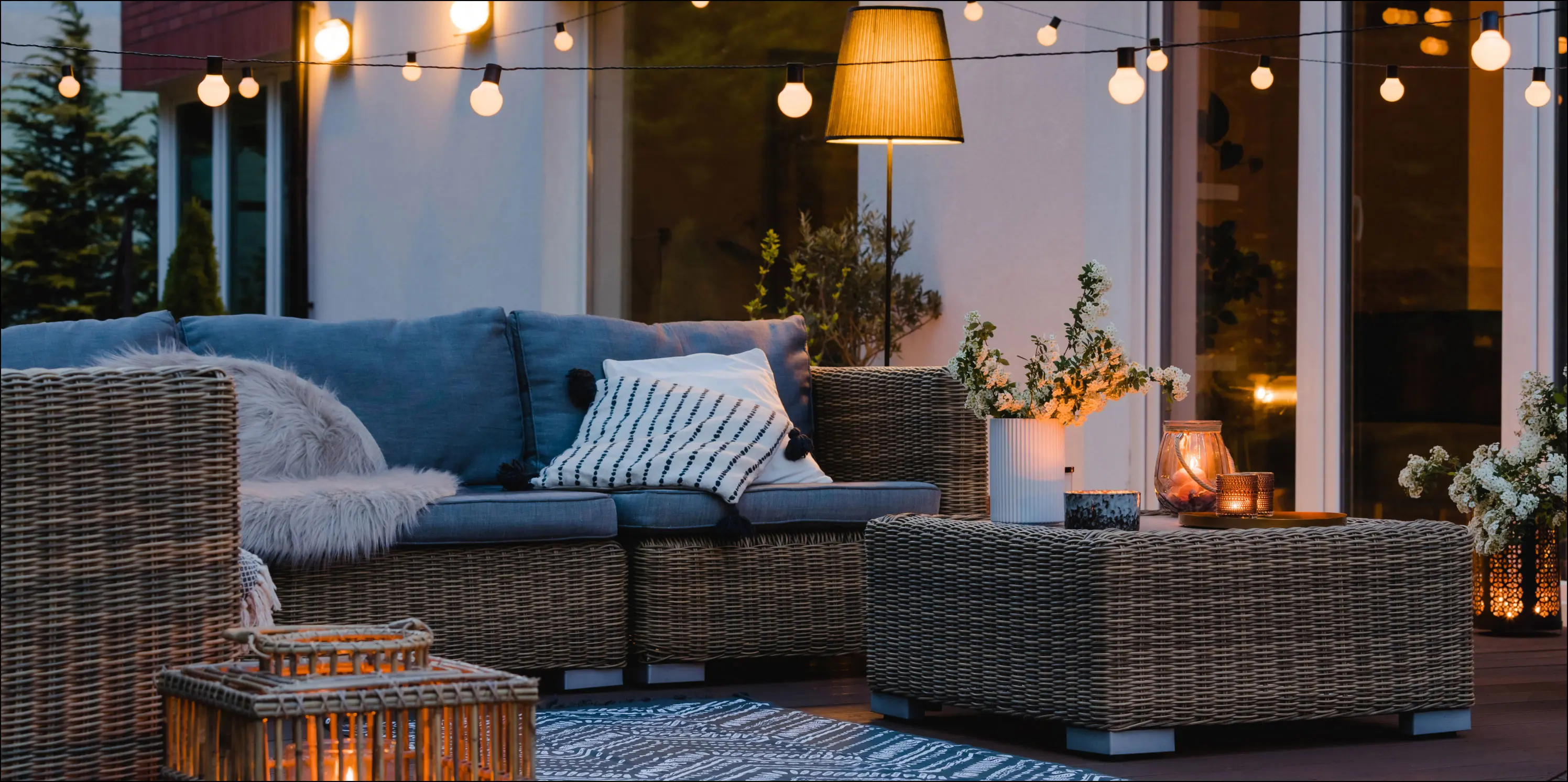 Een zithoek van rotan op een vlondervloer met sfeervolle verlichting in een tuin. 