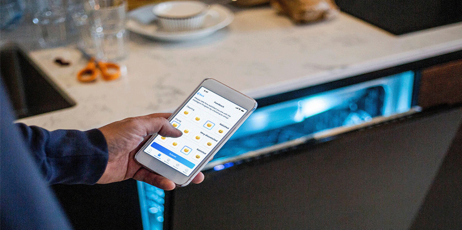 Hand houdt smartphone vast waarmee hij de vaatwasser op de achtergrond bedient. 