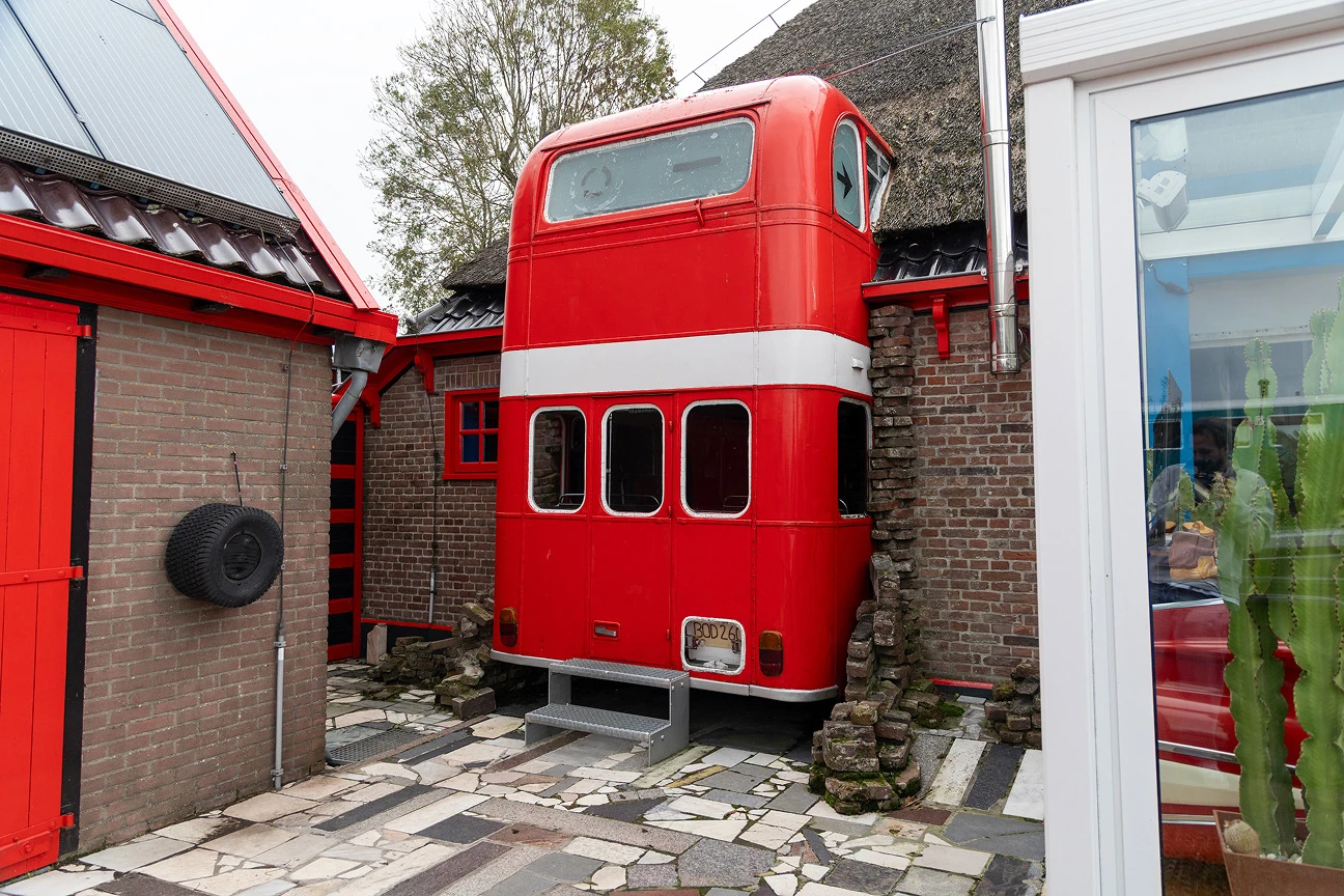 Een rode, Engelse dubbeldekkerbus lijkt door de gevel te zijn gereden. Alleen de achterzijde is zichtbaar, die deels uit de gevel en het dak steekt.