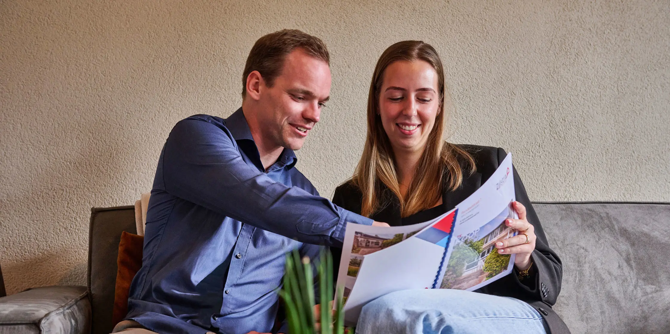Juliet en Robbert zitten op een lichtgrijze bank en bekijken een brochure van een huis. 