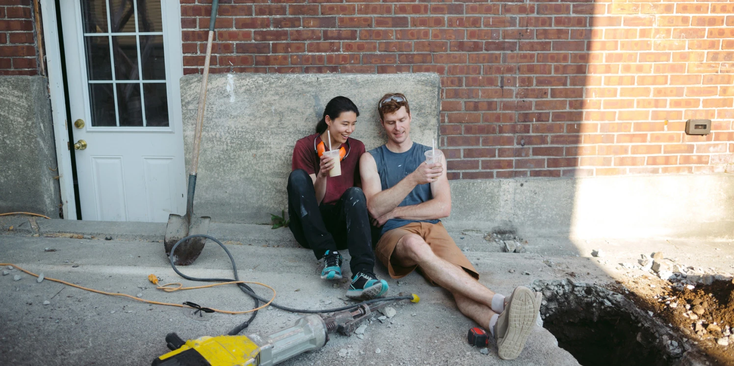 Een jong stel in kluskleren zit buiten op het beton te pauzeren met een smoothie in de hand.