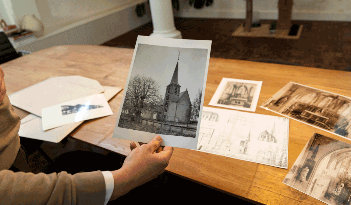 Een oude zwartwit-foto van de kerk, zoals deze vroeger was, vastgehouden door Rolf. Op de achtergrond liggen andere oude foto's en bouwtekeningen op de houten tafel.