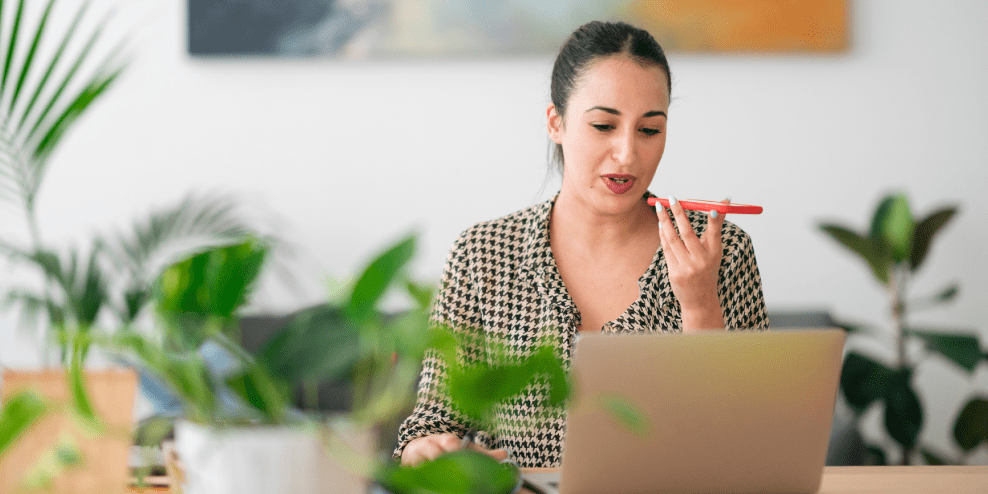 Vrouw aan bureau achter laptop met in haar hand een telefoon waar ze in praat.