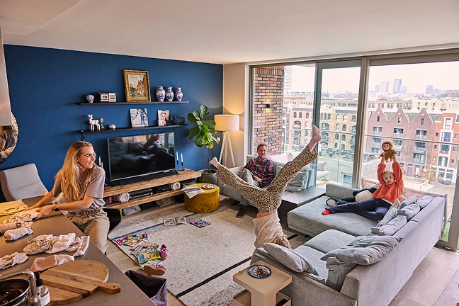Marga en Arjan met hun dochters in de woonkamer. Marga vouwt de was, oudste dochter maakt een handstand voor de bank. Arjan kijkt toe. Jongste dochter houdt knuffel omhoog.