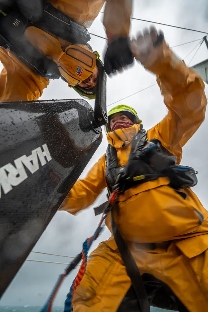 Low-angle view of a Clipper Race crew member in yellow foul-weather gear operating a winch during a sail manoeuvre on deck in offshore racing conditions.