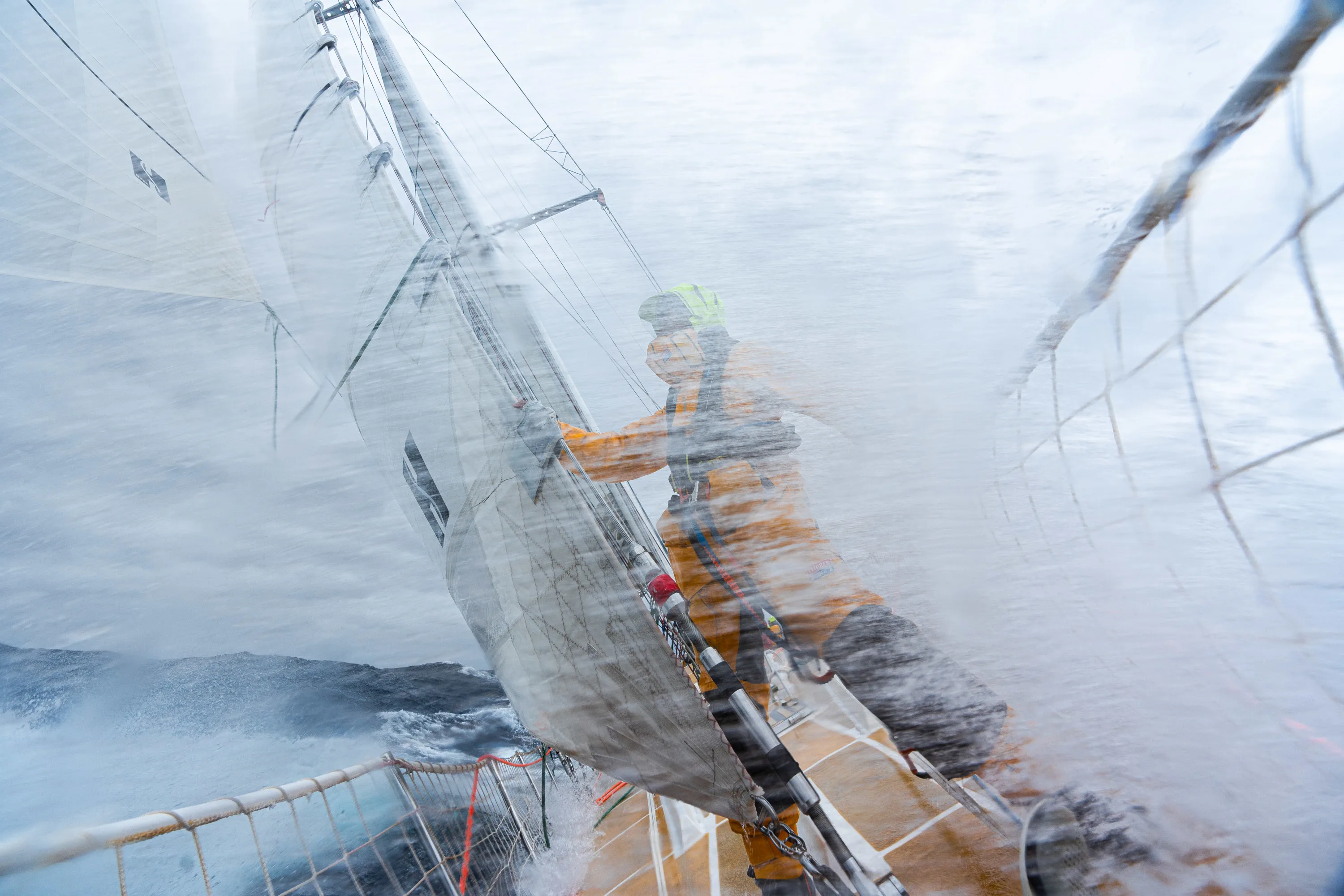 A crew member holds onto a sail as waves crash over the deck, capturing the intensity of ocean racing.