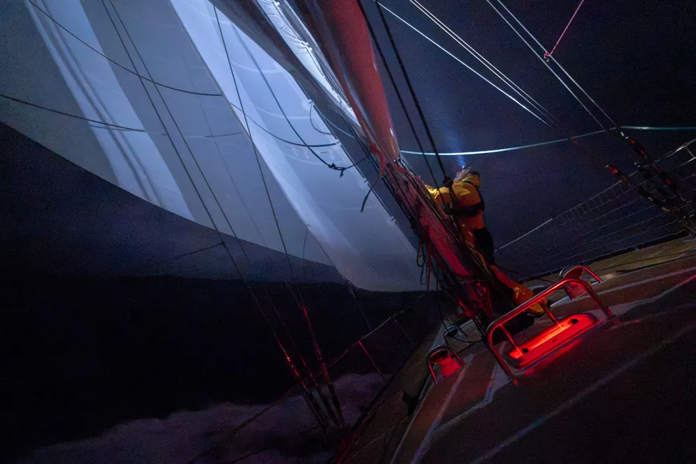 Clipper Race crew member working on the foredeck at night beneath illuminated sails, with red deck lighting and open ocean visible during an offshore race.