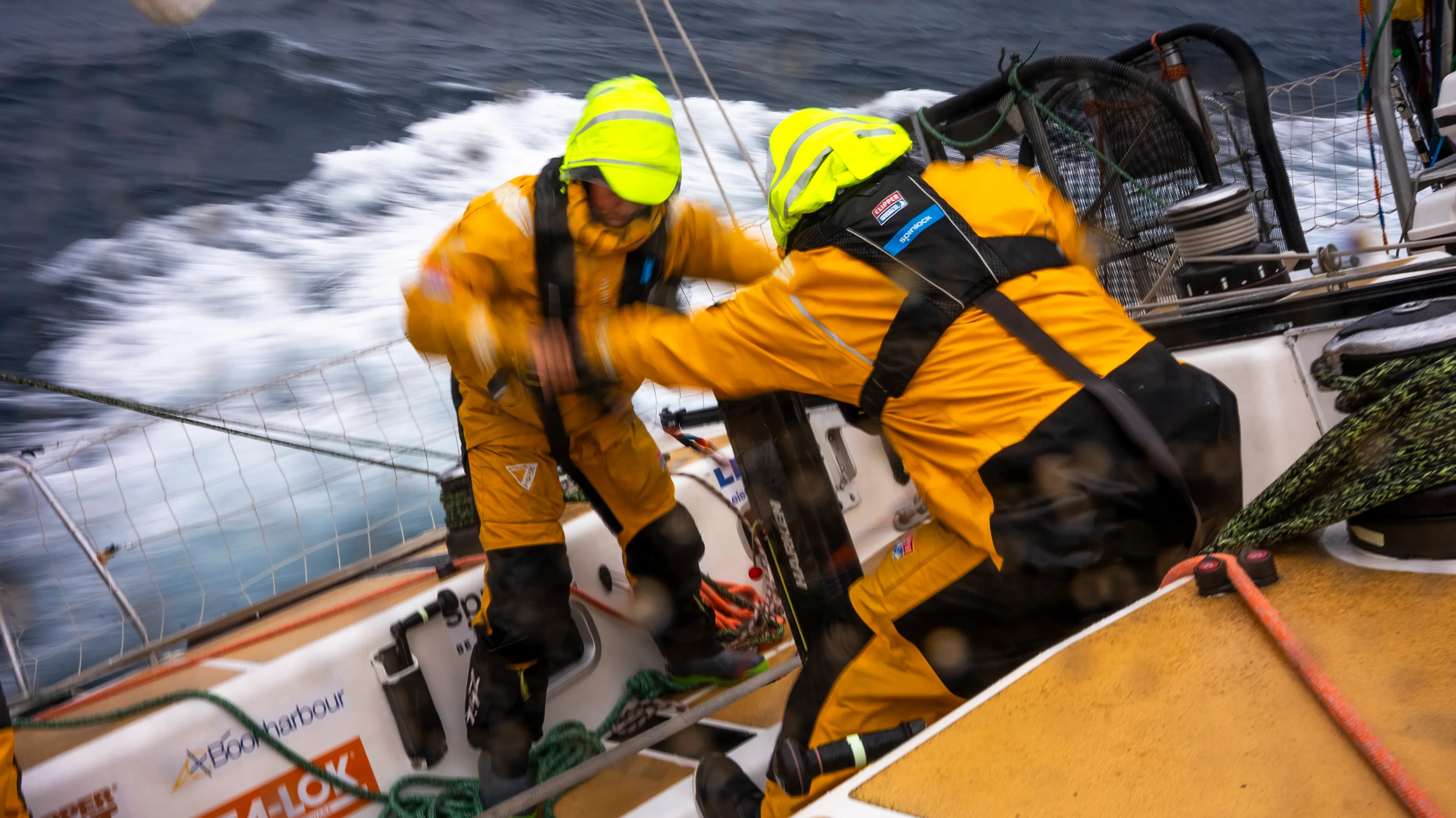 Two Clipper Race crew members in yellow foul-weather gear work together on deck during Leg 6, The Mighty Pacific, carrying out a sail manoeuvre as the yacht powers through rolling ocean swells.
