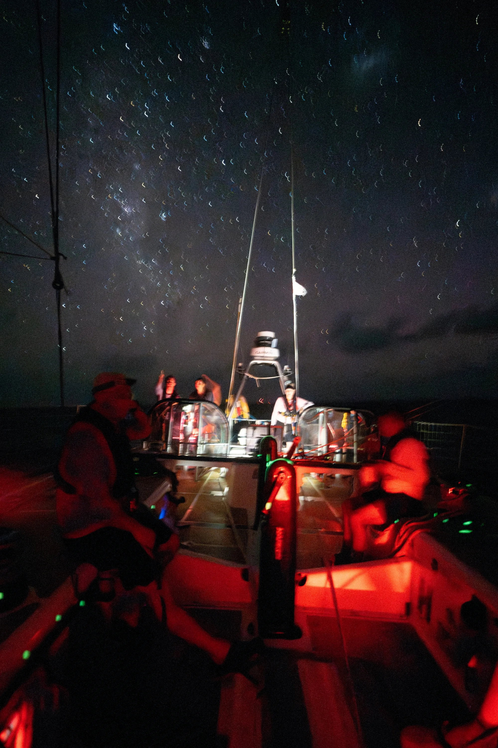 Clipper Race crew members on deck at night beneath a star-filled sky, illuminated by red deck lighting while sailing across the open ocean during an offshore race.