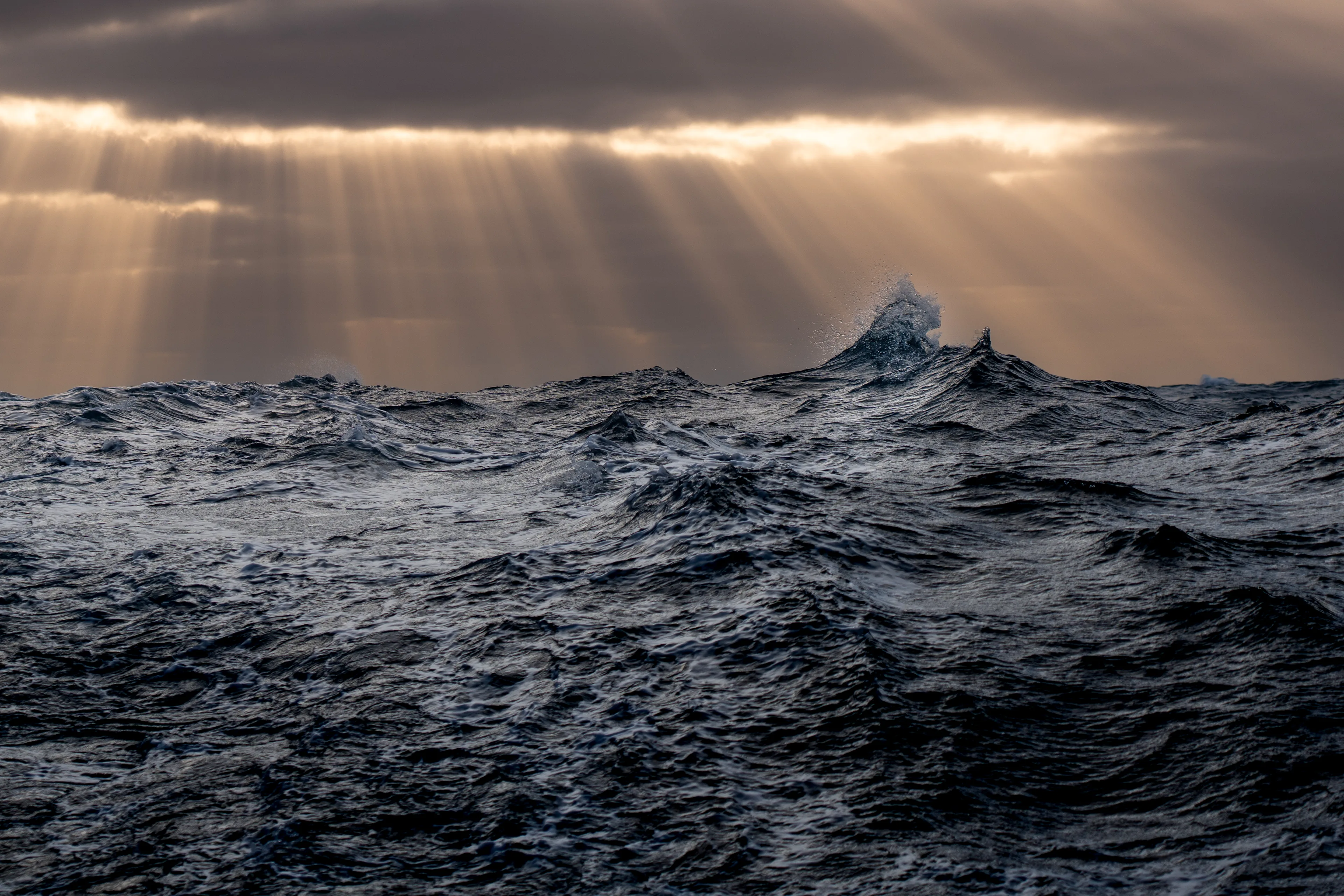 Dramatic ocean swells during Leg 6, The Mighty Pacific, with powerful waves rolling beneath sunbeams breaking through dark storm clouds on the horizon.