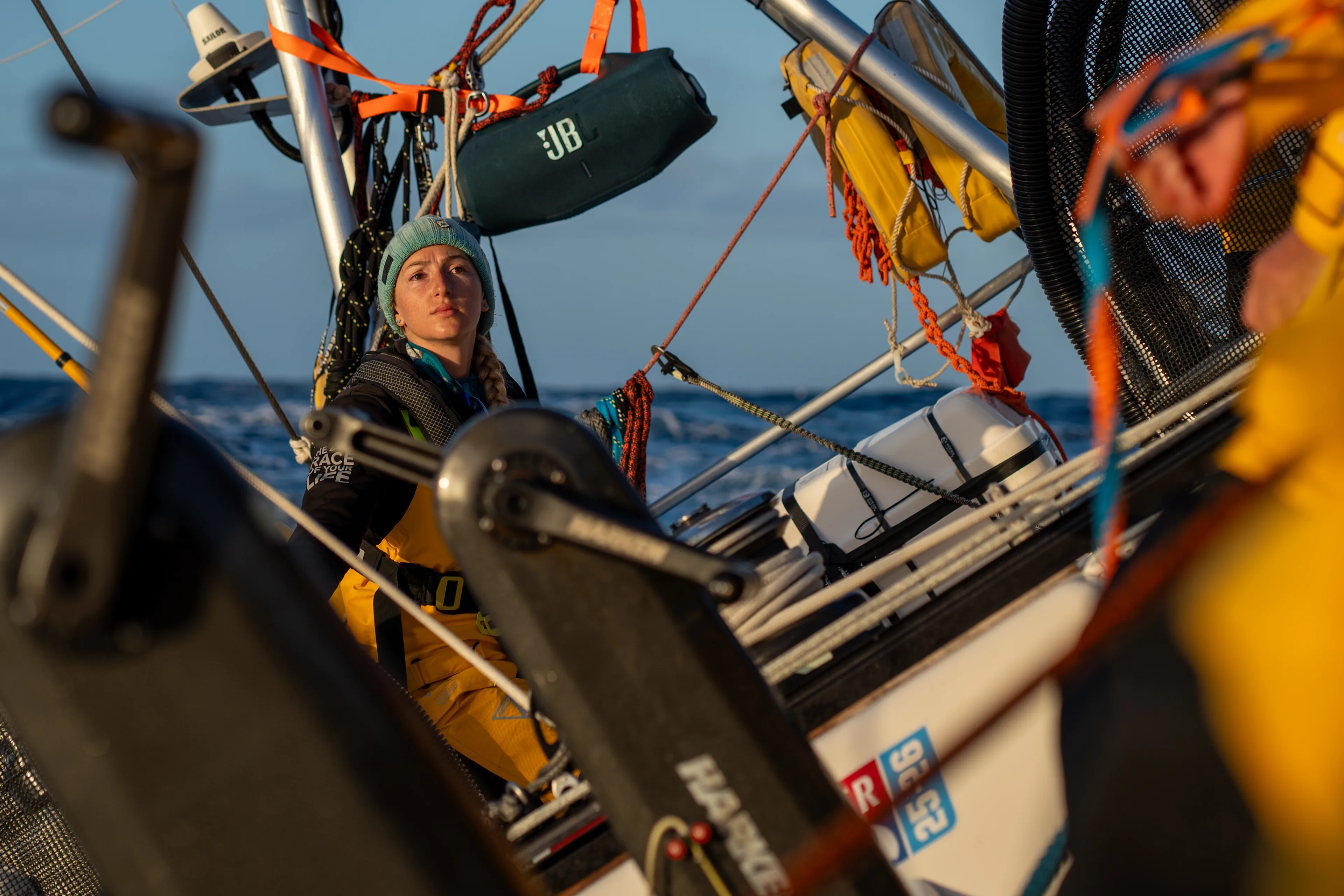 Clipper Race crew member seated near the helm during an offshore ocean crossing, surrounded by sailing equipment and rigging as the yacht races through open water.
