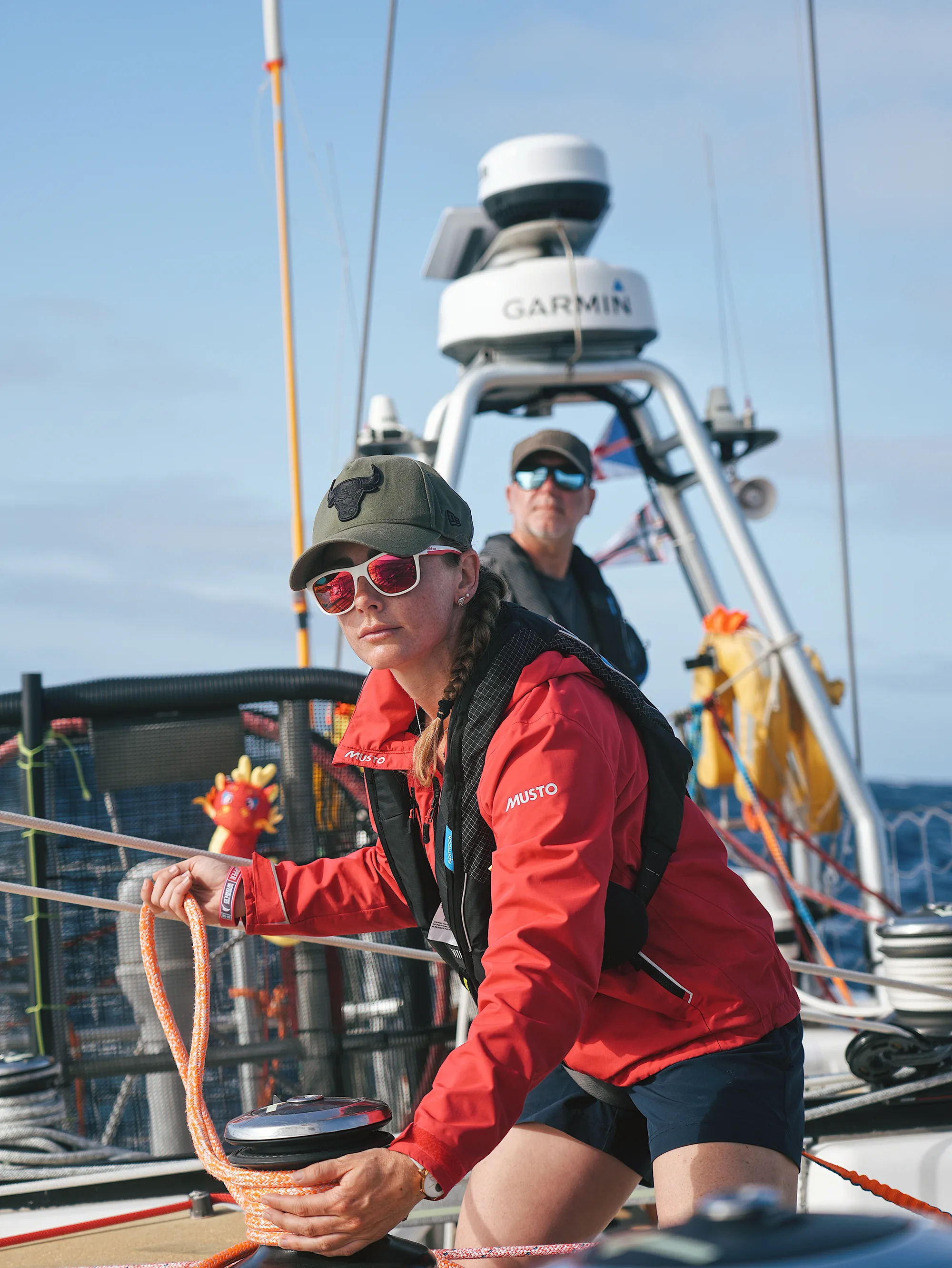 Clipper Race crew member wearing a red foul-weather jacket and sunglasses while handling sailing Marlow lines on deck, with another crew member and yacht equipment visible behind.