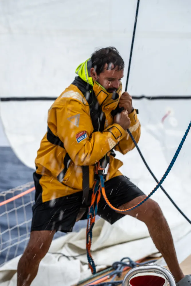 Clipper Race crew member in yellow foul-weather gear sweating sailing lines during a sail manoeuvre on deck in offshore ocean racing conditions.