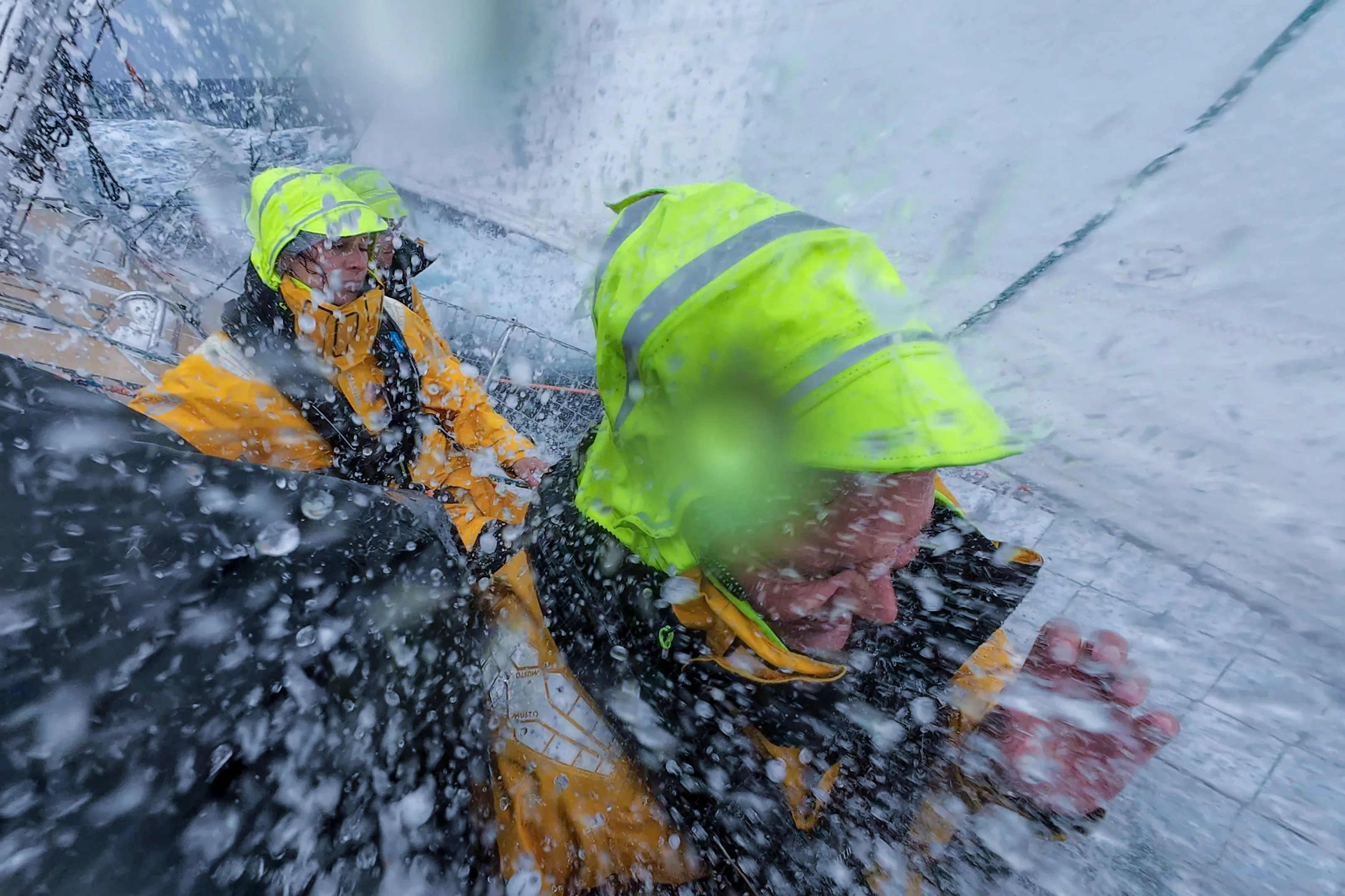 Two Clipper Race crew members in yellow foul-weather gear brace against heavy sea spray during Leg 6, The Mighty Pacific, as waves crash across the deck in challenging offshore conditions.