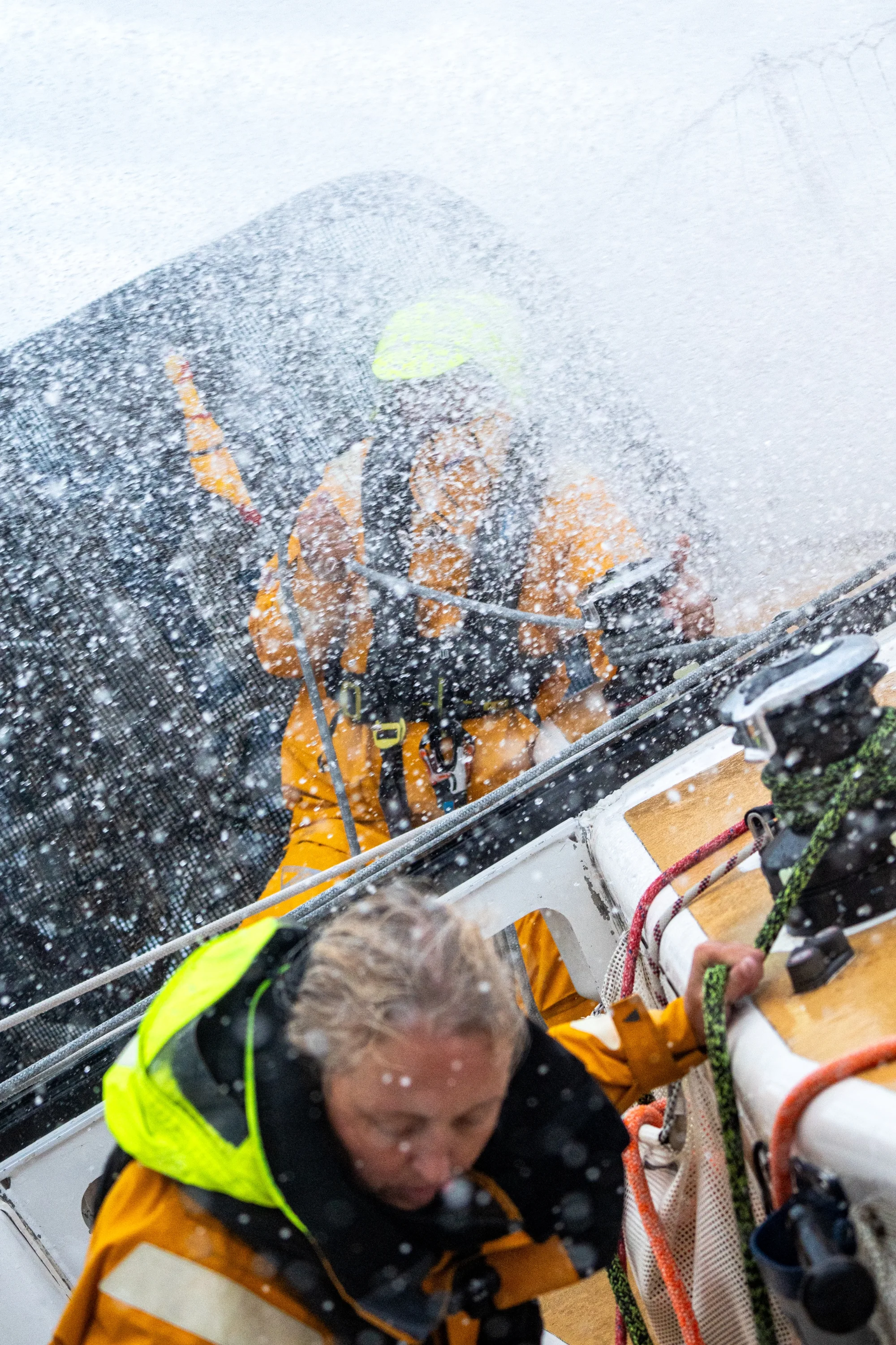 Clipper Race crew members in yellow foul-weather gear working on deck as heavy sea spray crashes over the yacht during challenging offshore racing conditions.