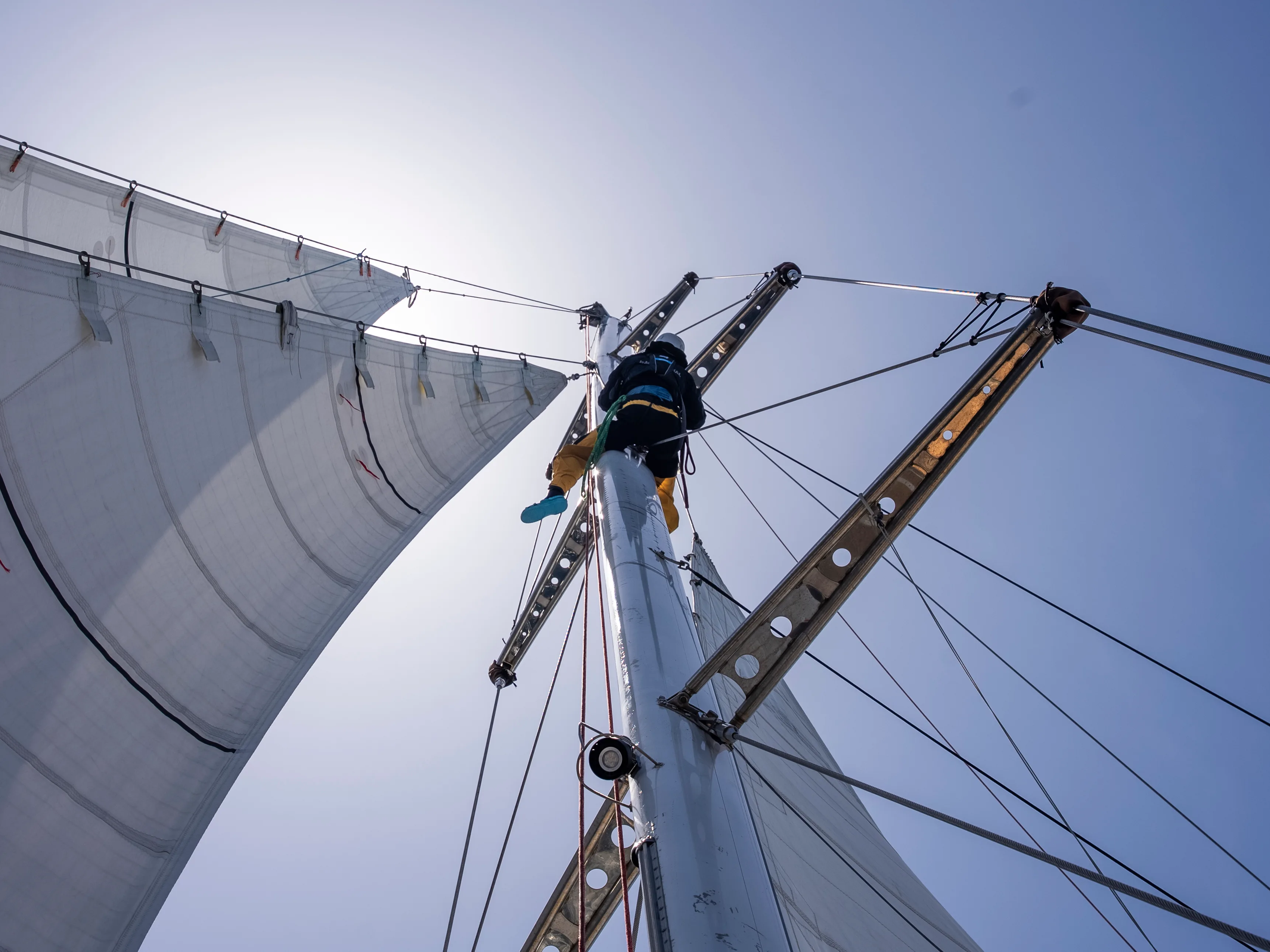 Clipper Race crew member climbing the mast while secured by safety lines during Leg 6, The Mighty Pacific, carrying out essential maintenance high above the deck under full sail.