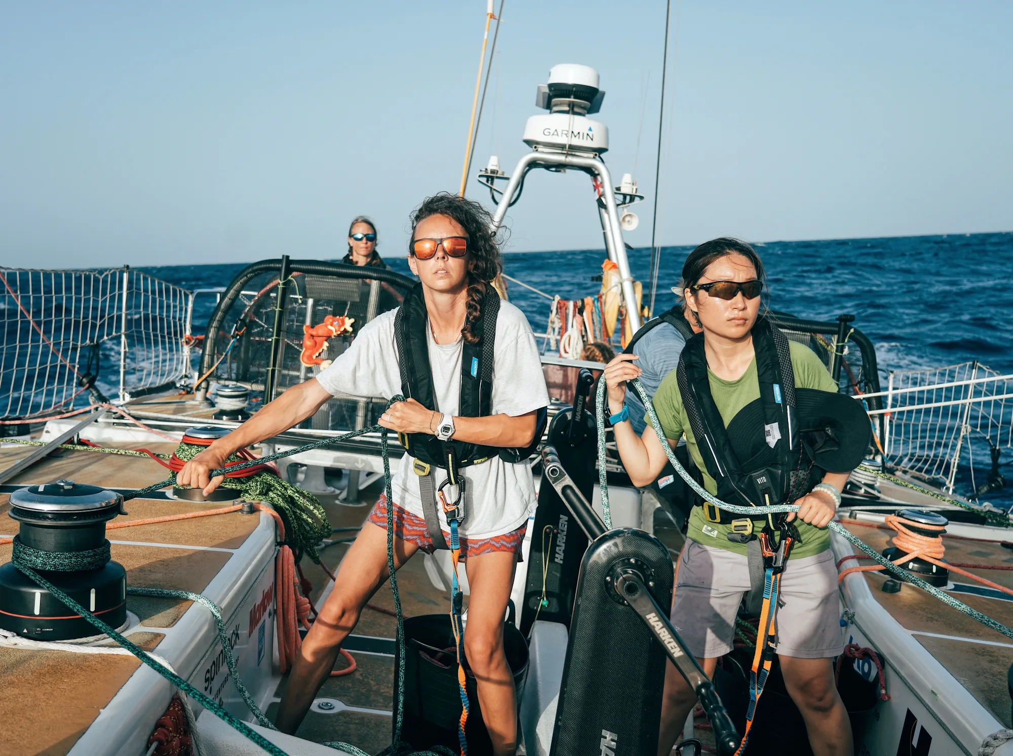 Two Clipper Race crew members steering and managing sail controls on deck during an offshore sailing race, with the open ocean stretching behind them.