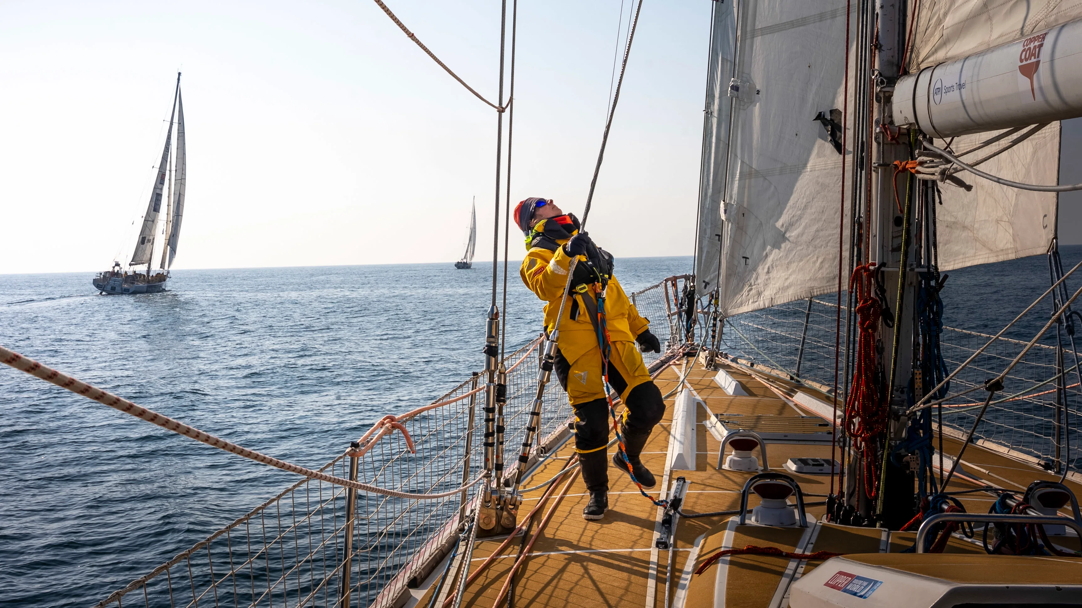 Clipper Race crew member on deck using binoculars to scan the horizon during Leg 6, The Mighty Pacific, with fellow race yachts visible in the distance across calm ocean waters.