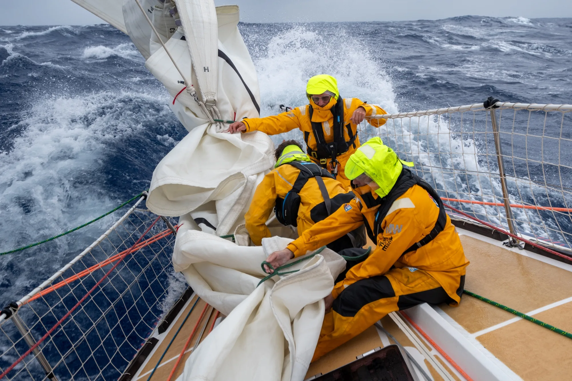 Clipper Race crew members in yellow foul-weather gear handling a sail on the foredeck as the yacht races through rough ocean conditions with large waves in the background.