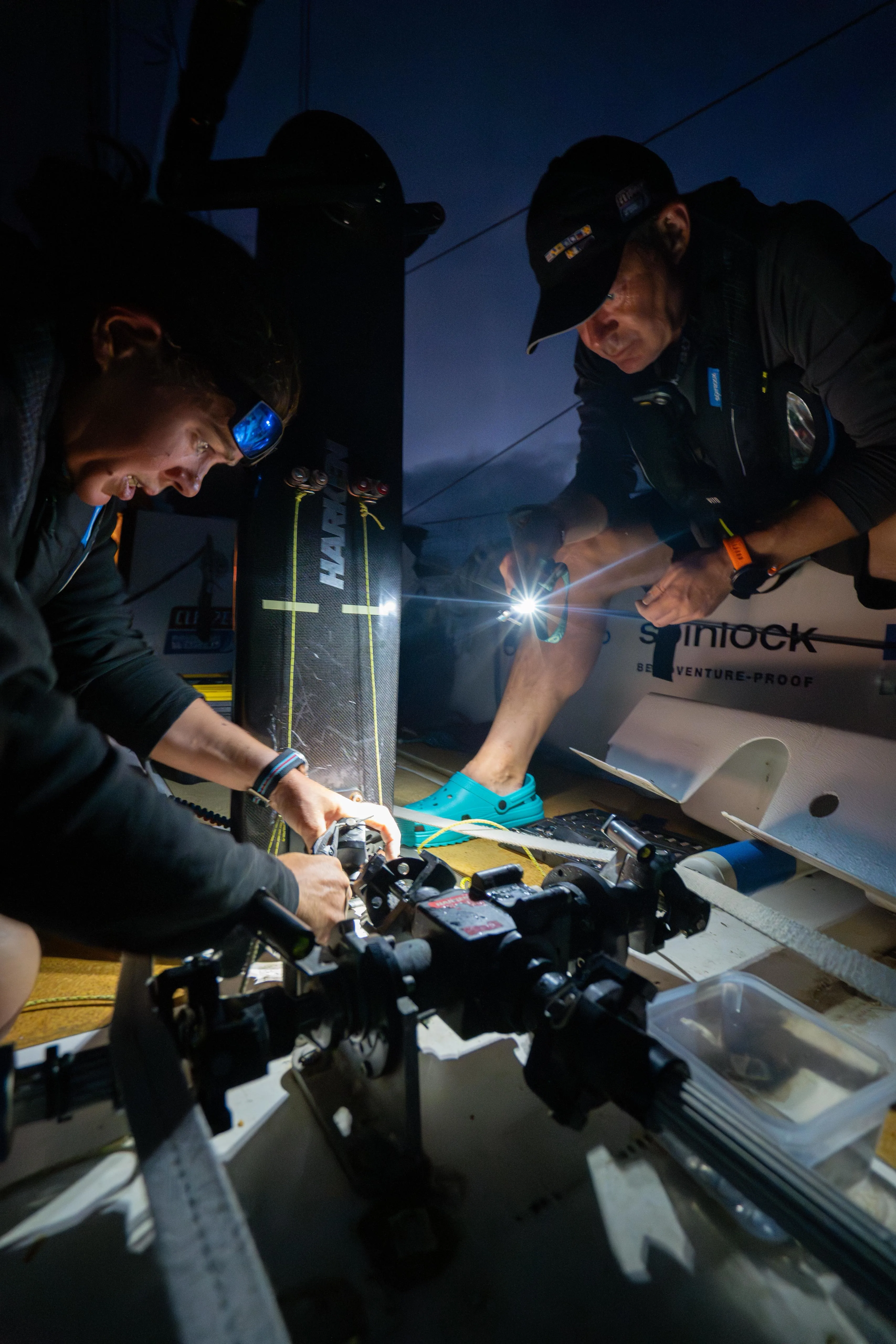 Clipper Race crew members using head torches to carry out maintenance and repairs on deck during a night watch on an ocean racing yacht.