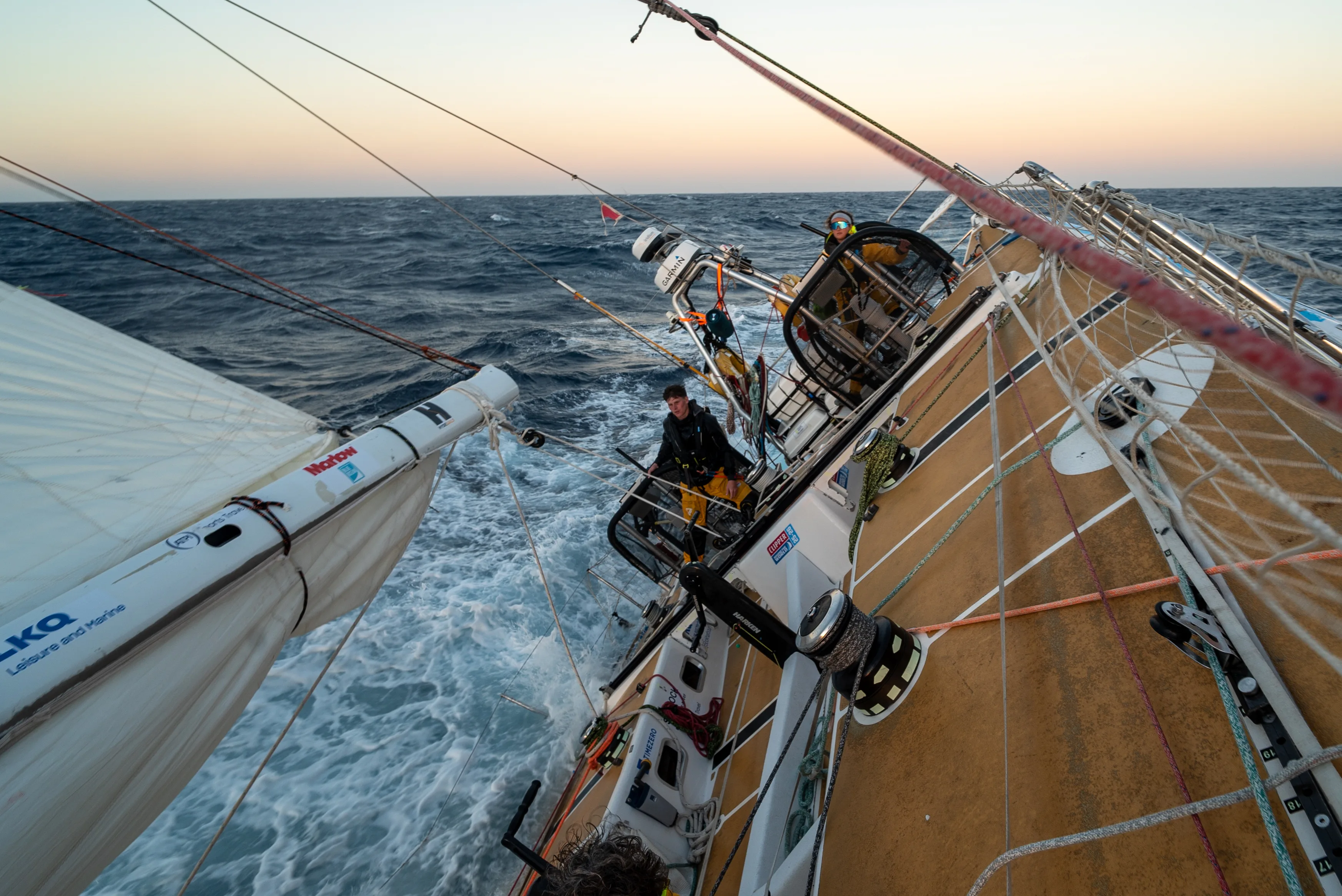 Clipper Race yacht sailing at an angle through rough seas at sunset, with crew members working on deck during an offshore ocean race.