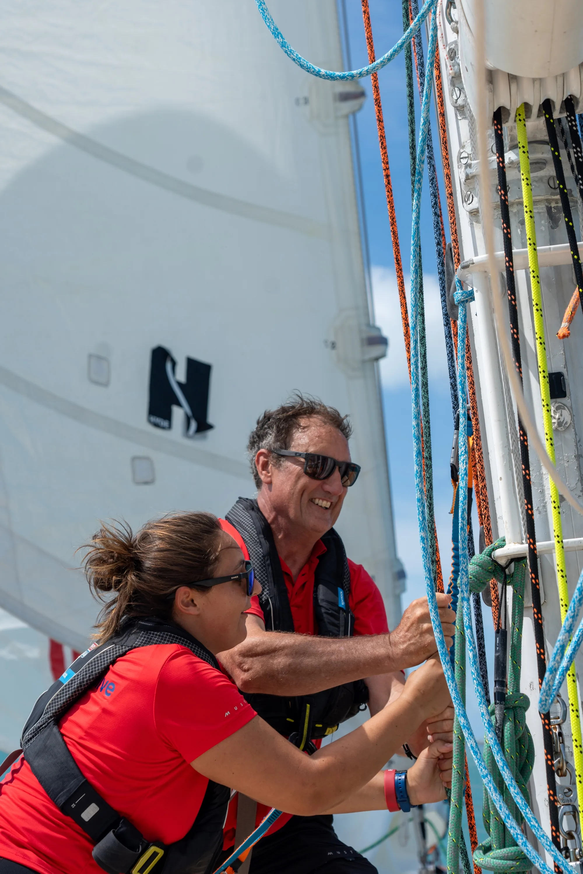 Two Clipper Race crew members adjusting sailing lines and rigging on deck during an offshore ocean race, working together to optimise yacht performance under sail.