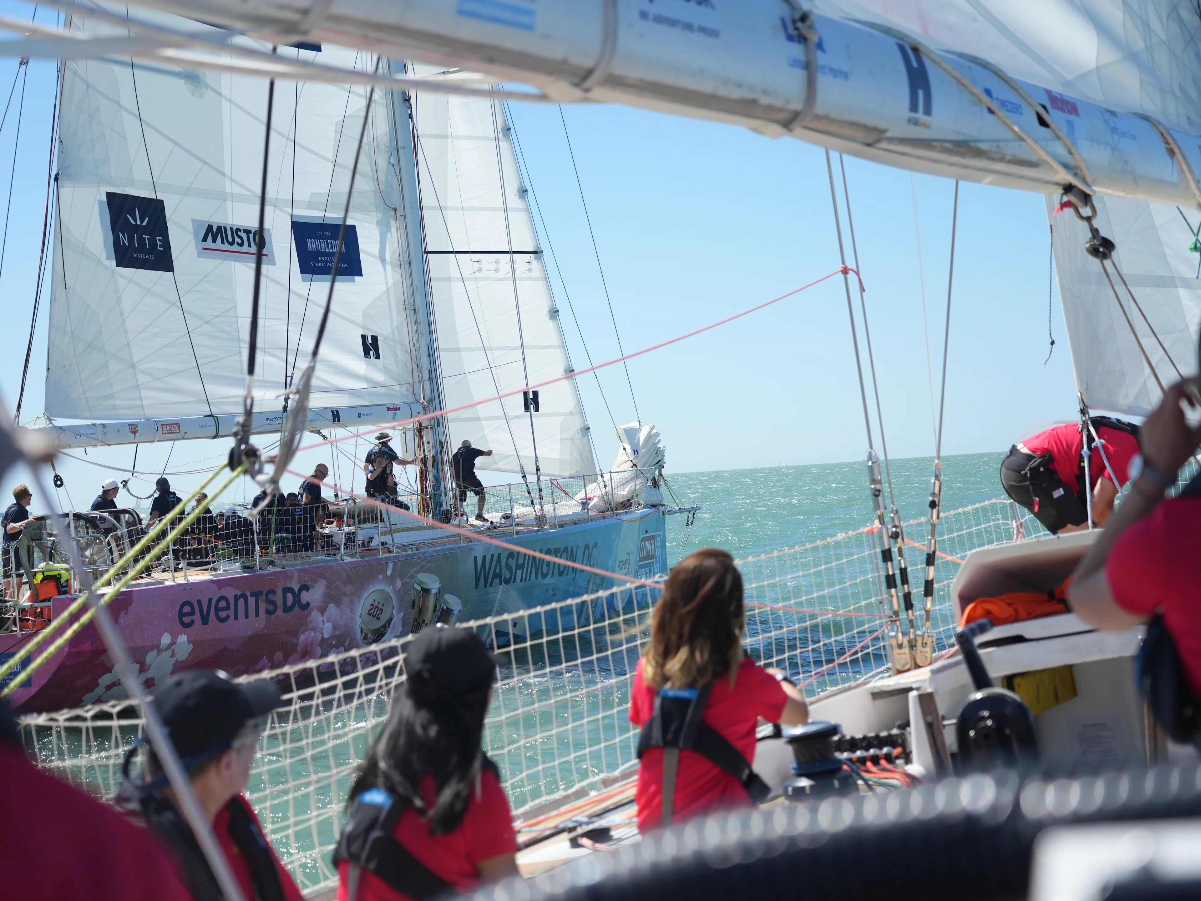 View from the deck of a Clipper Race yacht as the Washington, DC team sails alongside Qingdao during a race start, with crews visible on both boats.