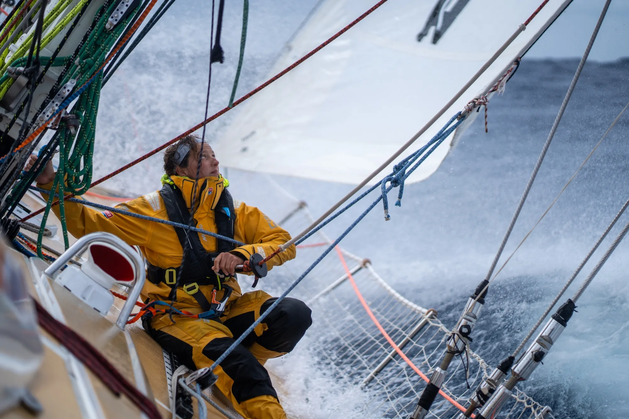Clipper Race crew member in yellow foul-weather gear adjusting sail controls on deck as waves crash over the yacht during rough offshore racing conditions.