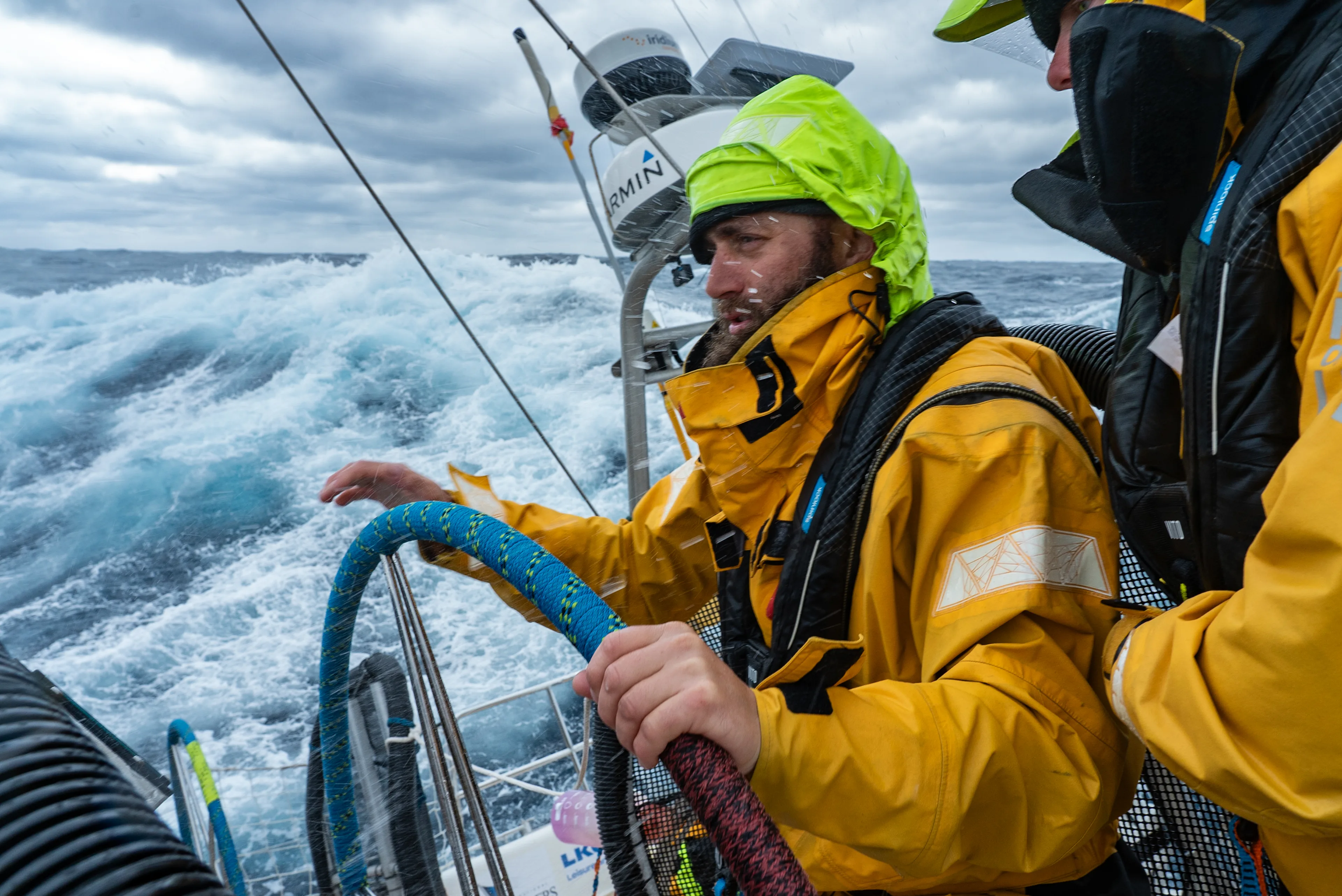 Clipper Race crew member at the helm during Leg 6, The Mighty Pacific, steering through large ocean swells in challenging offshore conditions while another crew member stands watch nearby.