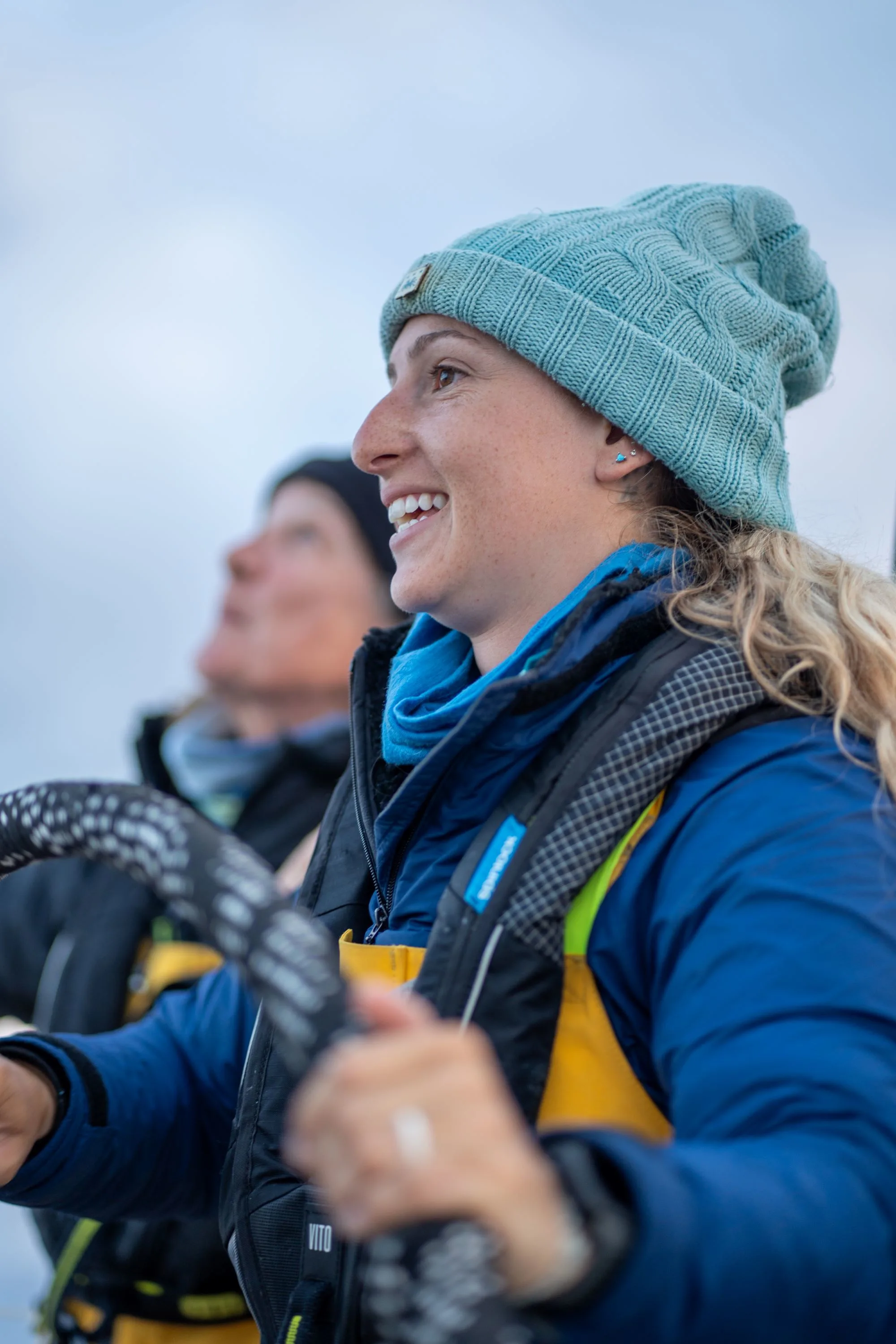Smiling Clipper Race crew member wearing a blue beanie and lifejacket while steering the yacht during an offshore ocean sailing race.