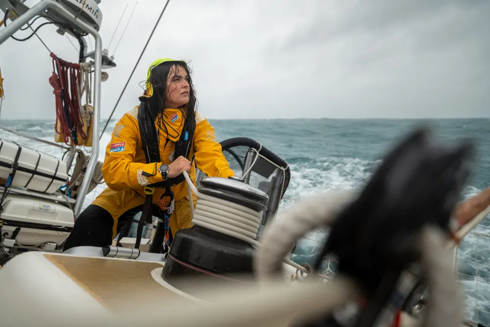 Diana, First Mate of the UNICEF Clipper Race team, steering the yacht in foul-weather gear during an offshore ocean race, with waves and overcast skies in the background.