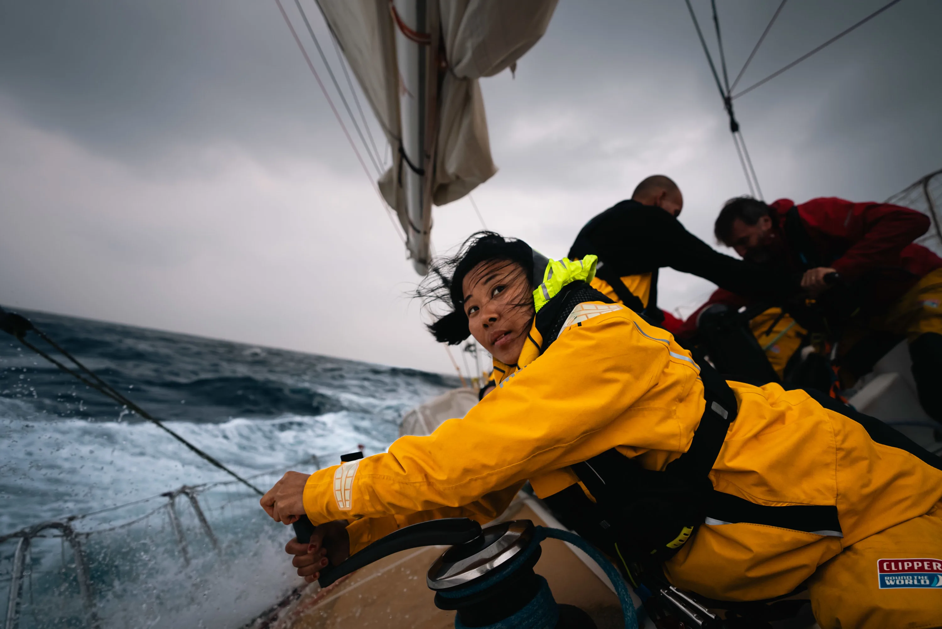 A woman grips a winch handle on a sailing boat, bracing herself against the turbulent ocean waves under a dark, cloudy sky. The image captures the intensity and teamwork required for ocean racing in challenging conditions.