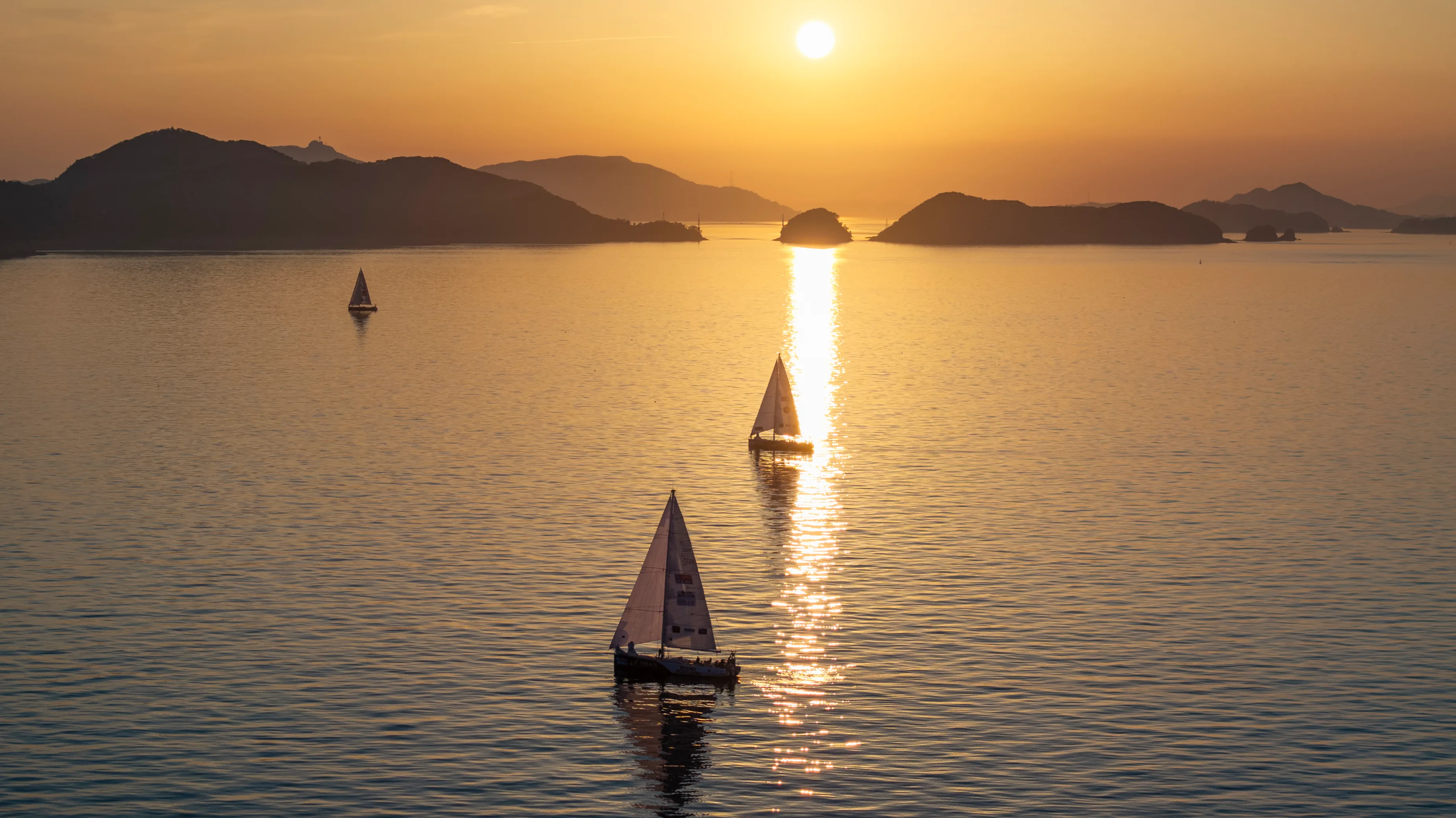 Clipper Race yachts sailing across calm waters at sunset during Leg 6, The Mighty Pacific, with golden light reflecting across the ocean and mountainous islands silhouetted on the horizon.