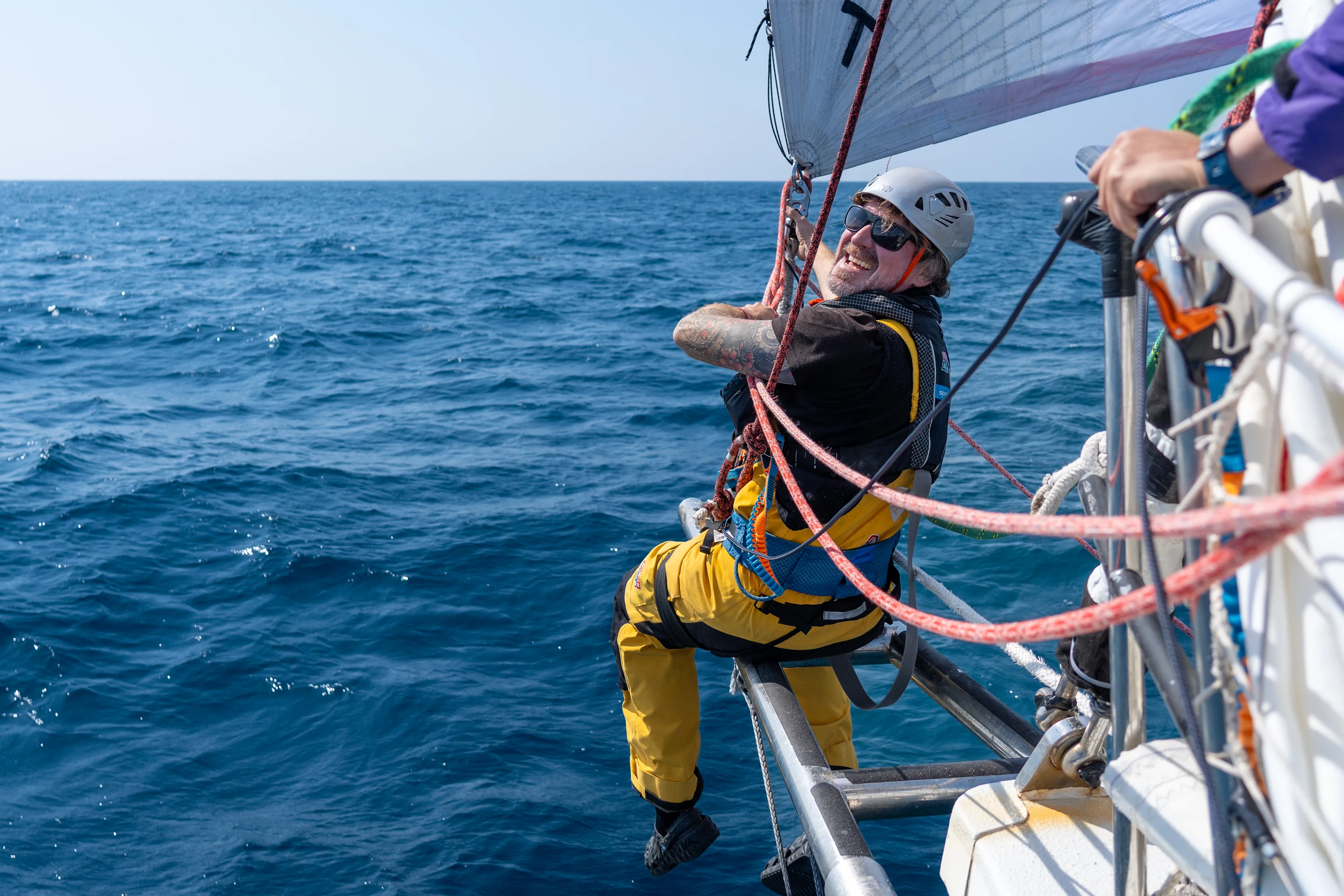 Clipper Race crew member suspended from the side of the yacht while secured by safety lines during Leg 6, The Mighty Pacific, carrying out maintenance above the open ocean.