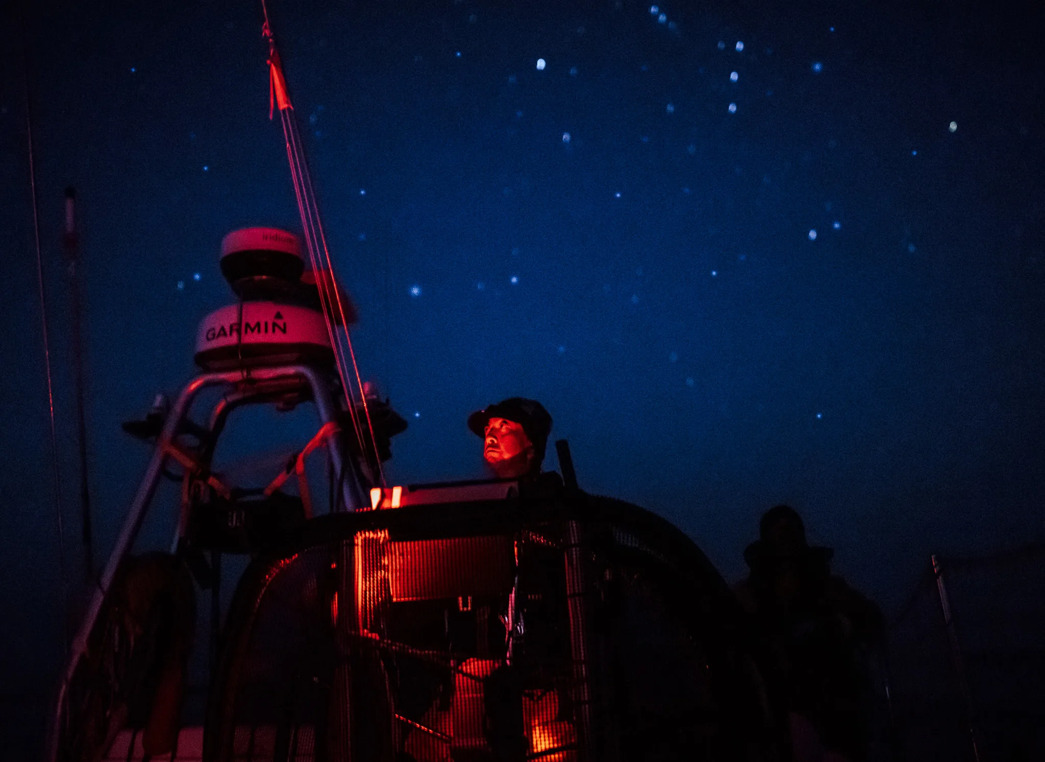 Clipper Race crew member standing on deck under a star-filled night sky, illuminated by red navigation lighting during an offshore ocean race.