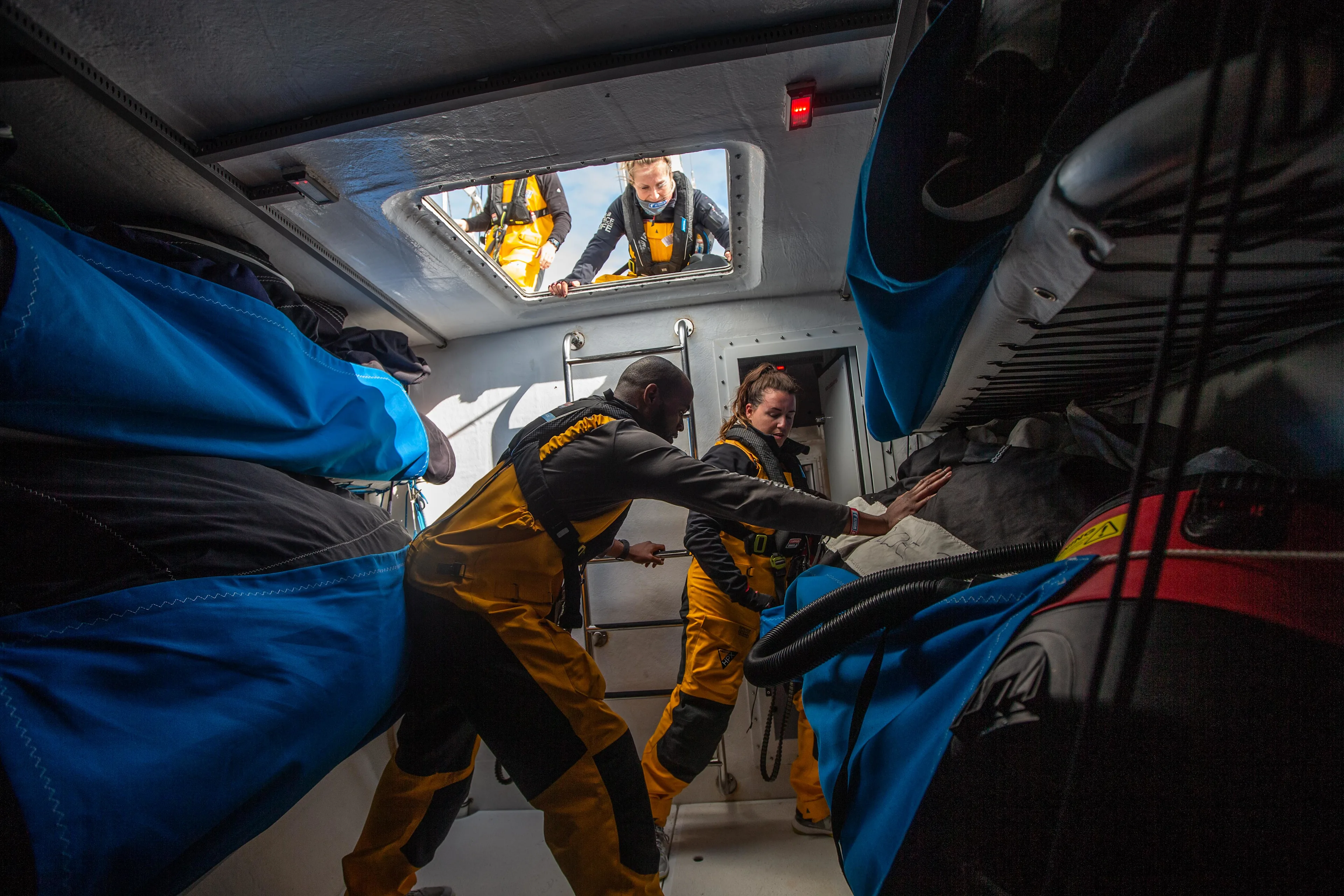 Race Crew inside the tight quarters of a sailboat. One crew member climbs down a ladder from an open hatch above, while two others organize equipment in the bunk area. Blue storage bags and gear are secured on either side of the narrow space, highlighting the cramped and functional setup needed for an ocean race.