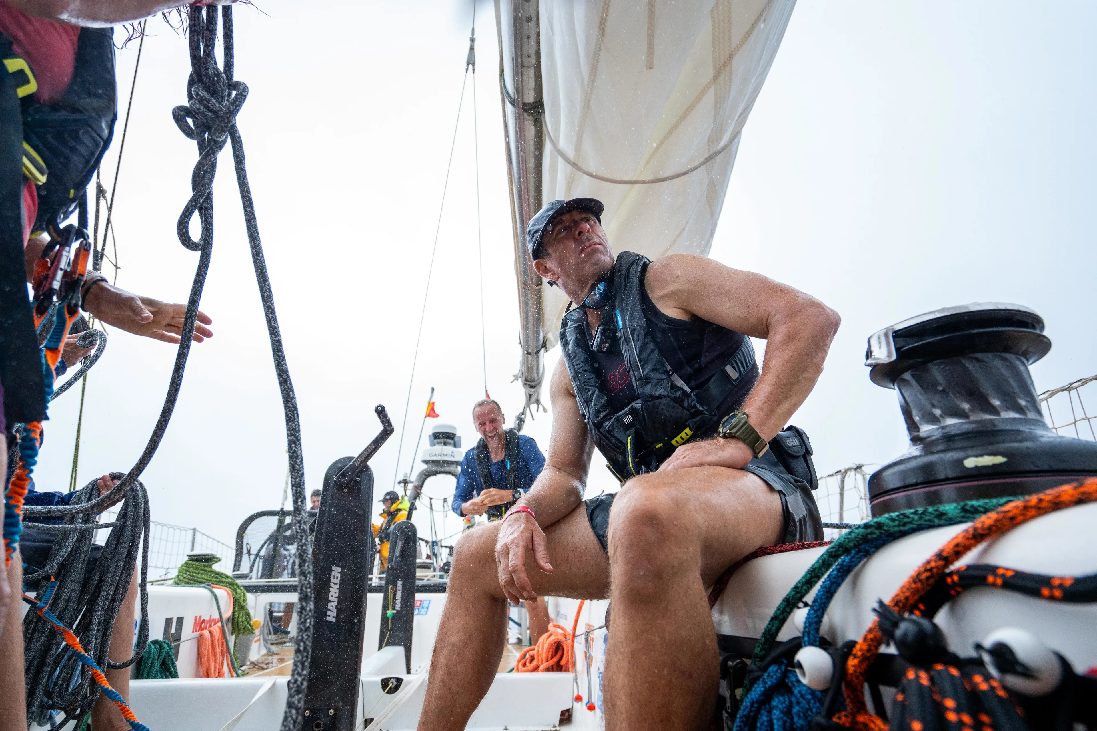 Clipper Race crew member seated on deck beside sailing equipment and winches during an offshore ocean race, with fellow crew visible in the background.
