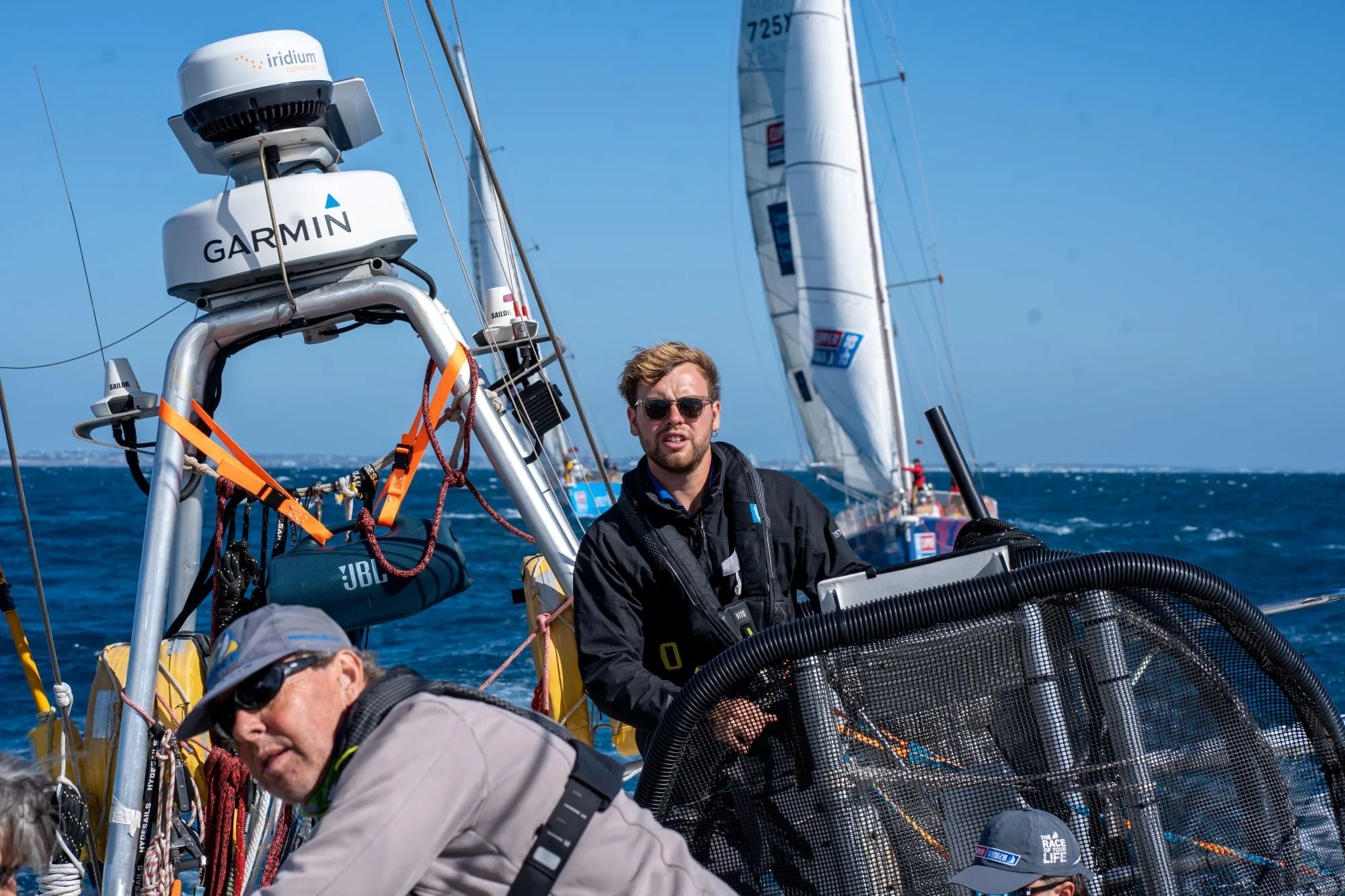 Clipper Race Skipper Oliver Irvine steering the yacht from the helm during an offshore ocean race, with crew members and sailing equipment visible on deck.