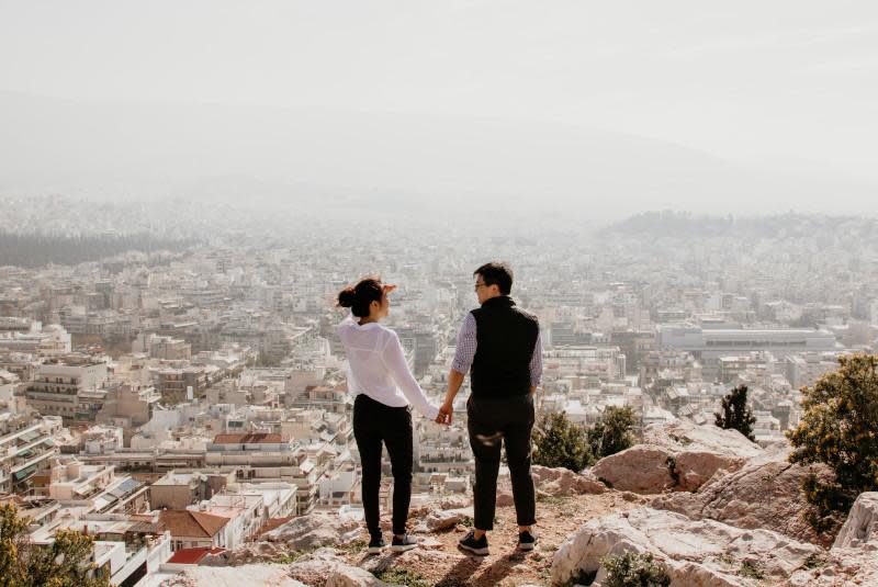 A couple standing on a rocky hillside viewpoint, looking out over a vast cityscape below. The person on the left wears a white top and dark pants and is pointing at something in the distance, while the person on the right, dressed in a checked shirt and dark vest, stands close beside them. They are viewed from behind, holding hands as they gaze at the sprawling white buildings of what appears to be Athens, Greece. The city stretches out across the valley and surrounding hills, slightly hazy in the distance. The scene captures a moment of connection while travelers take in a breathtaking panoramic vista.