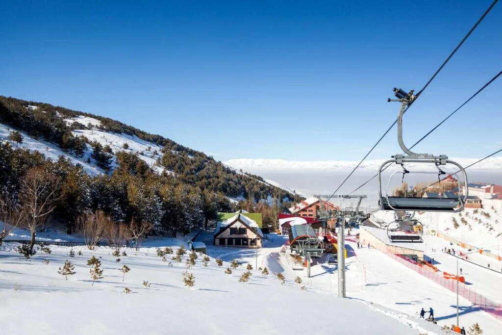 A chairlift rises above a Turkish ski resort with a wooden lodge, snowy slopes, and a hazy valley in the distance