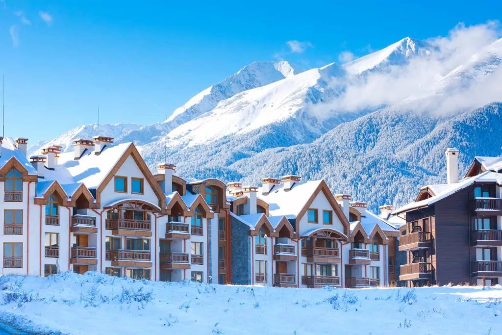 Snow-covered chalet-style buildings line a ski resort street in Bansko, Bulgaria, with mountains rising behind