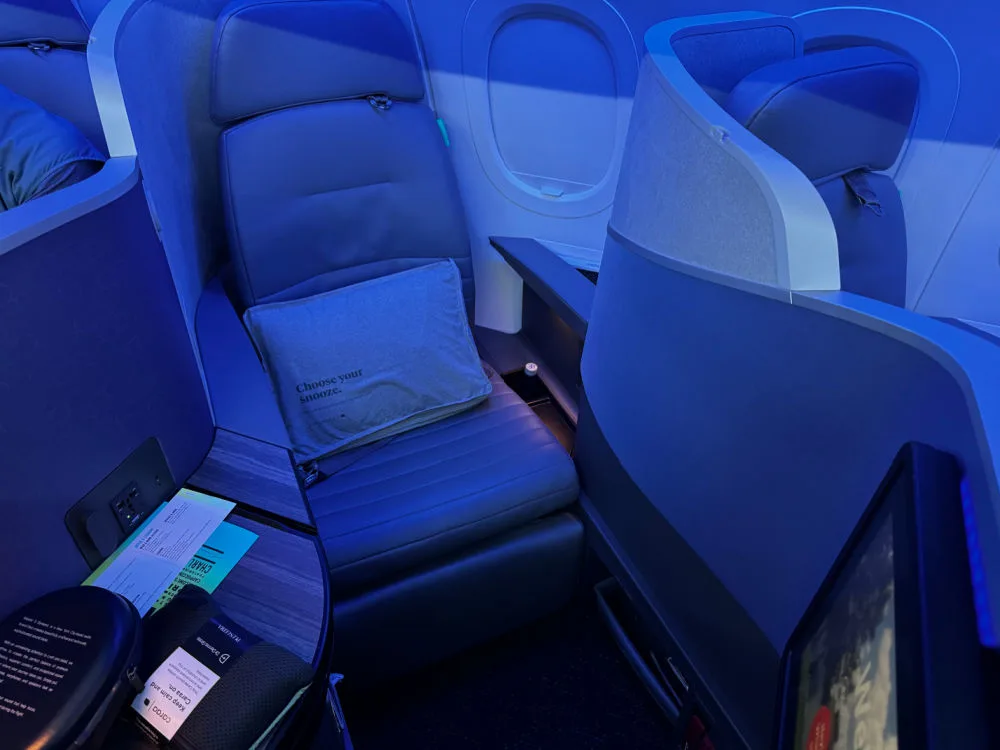 JetBlue Mint business class suite with a lie-flat seat and a pillow reading 'Choose your snooze' under blue mood lighting